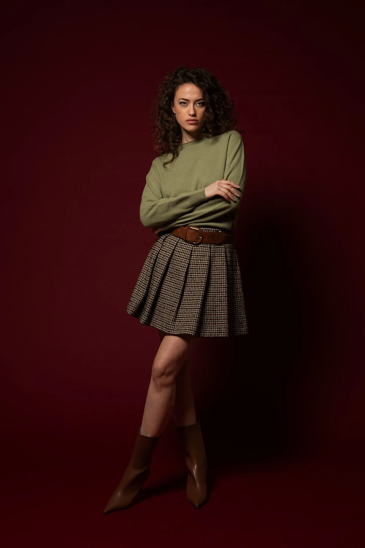 Donna con vestito a quadri, maglia verde e stivali marroni, posa con braccia incrociate, sfondo rosso scuro.