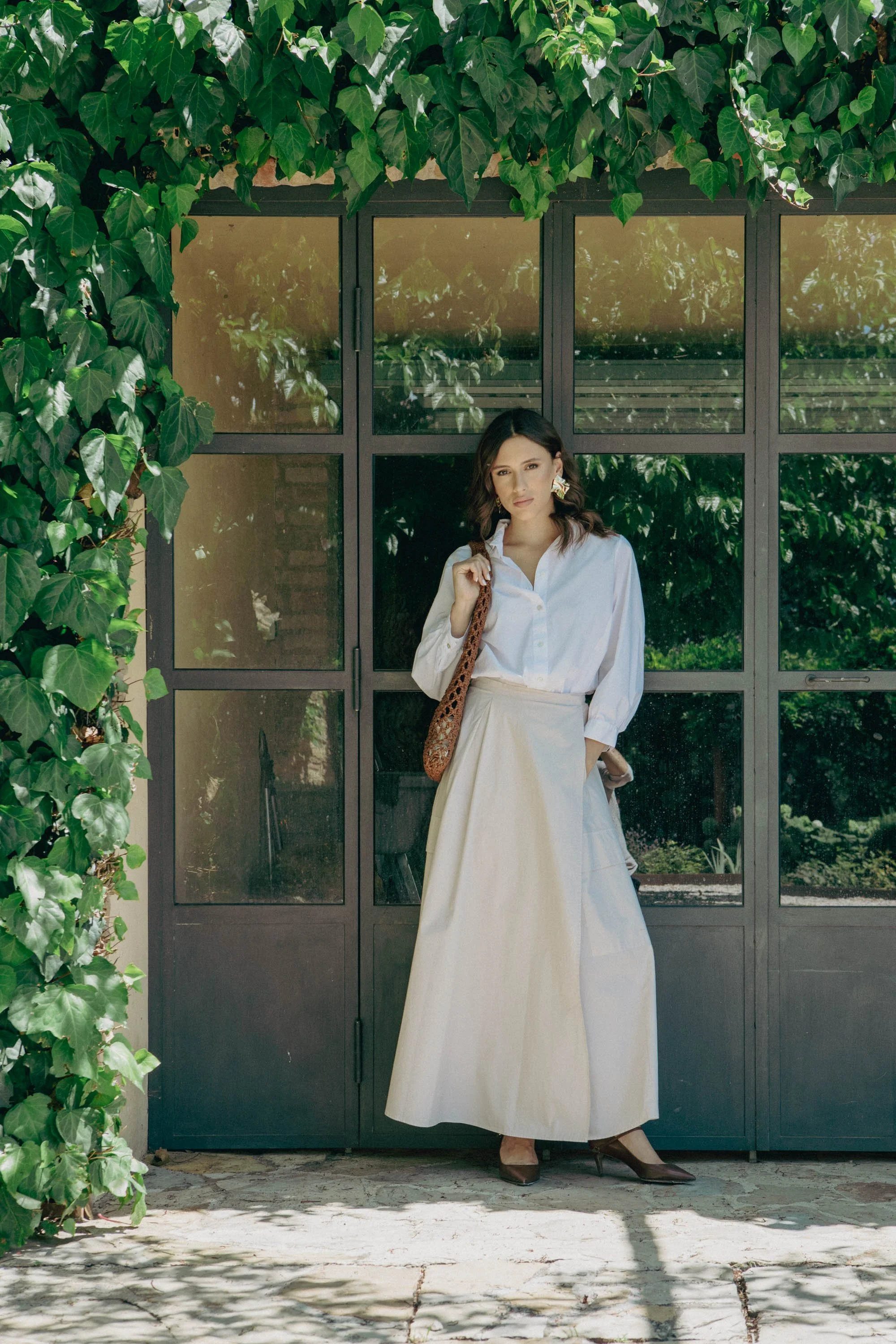 Una donna vestita di bianco con pantaloni lunghi e scarpe eleganti si trova davanti a una porta con vetri. La porta è circondata da foglie di edera verde sulle parti superiori e a sinistra. La donna porta una borsa a tracolla marrone con motivo leopa