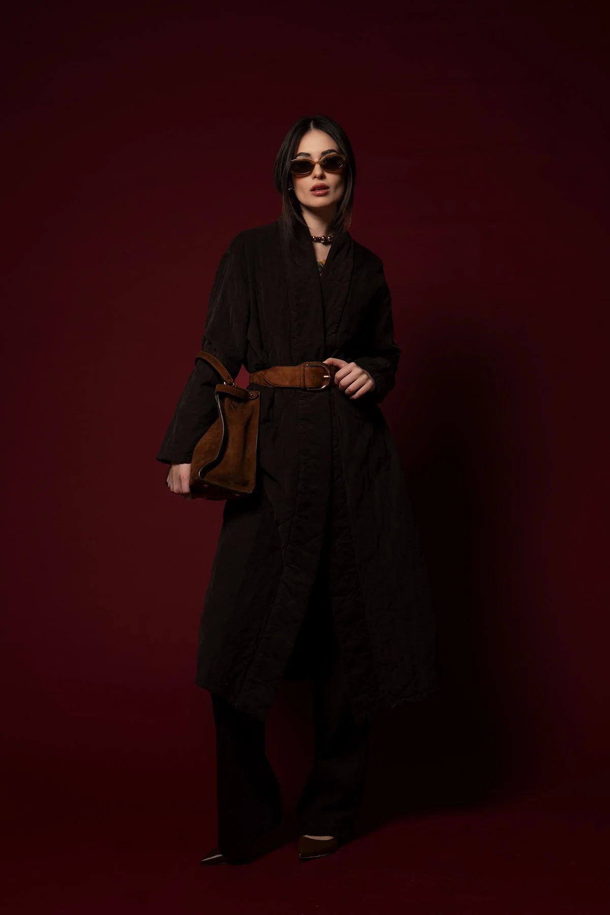 Una donna elegante con occhiali da sole neri, indossa un lungo vestito nero, una cintura marrone e una borsa in pelle marrone, posa davanti a uno sfondo rosso scuro.