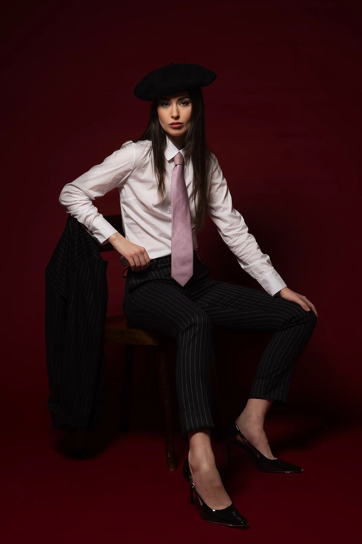 Donna seduta su una sedia con sfondo rosso, indossa una camicia bianca, cravatta rosa, pantaloni a pinstripe neri e tacchi neri, con un cappello nero.