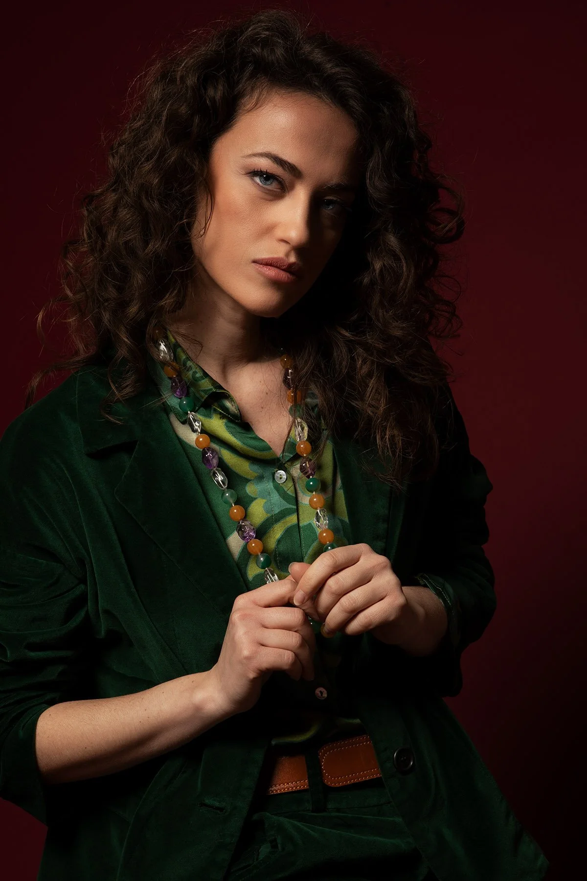ritratto di una donna con capelli ricci, vestita con giacca verde e collana di perline colorate, sfondo sfumato rosso