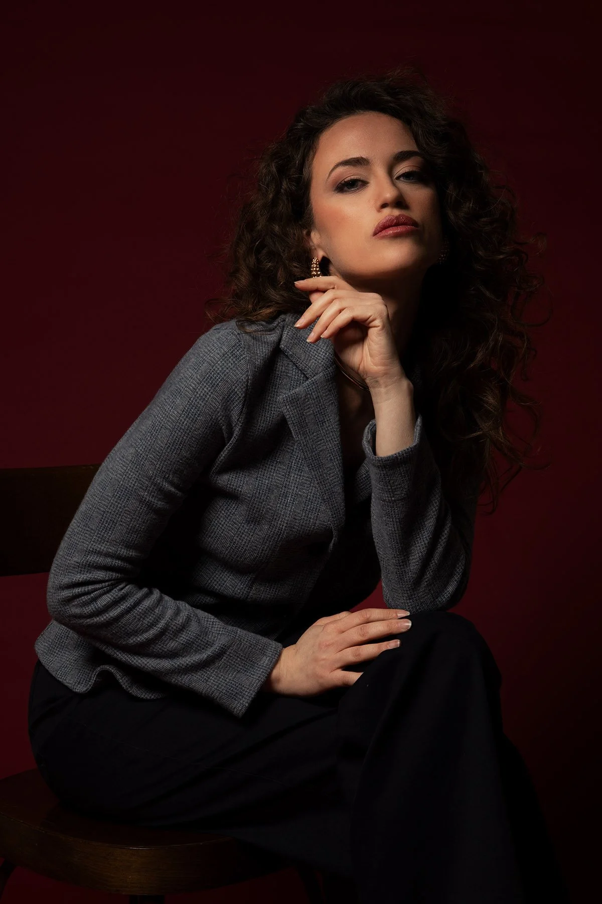 Una donna con capelli ricci e trucco marcato, indossa un abito grigio a quadri e orecchini dorati, mentre si siede su una sedia di legno scuro, posando con un'espressione sicura sul volto contro uno sfondo rosso scuro.