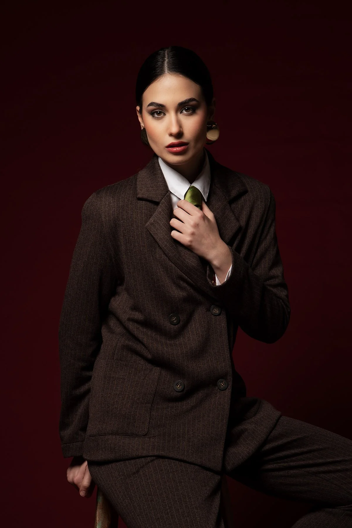 Una donna vestita elegantemente in completo marrone scuro con una camicia bianca, sfondo rosso scuro.