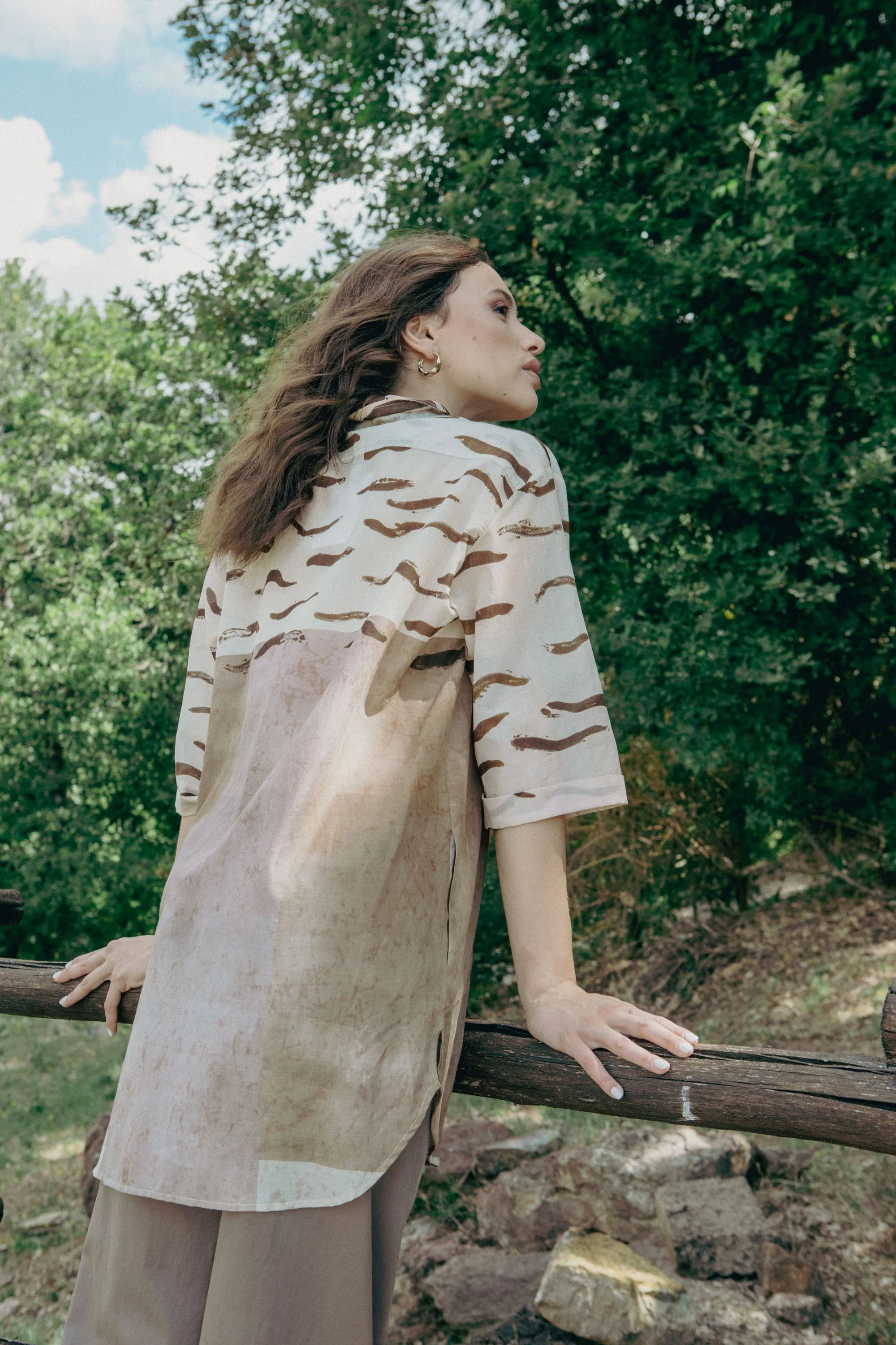Una donna con i capelli castani e corti, indossa un cappotto beige con motivo a strisce marroni, si appoggia a un albero in un ambiente naturale con alberi e cielo nuvoloso.