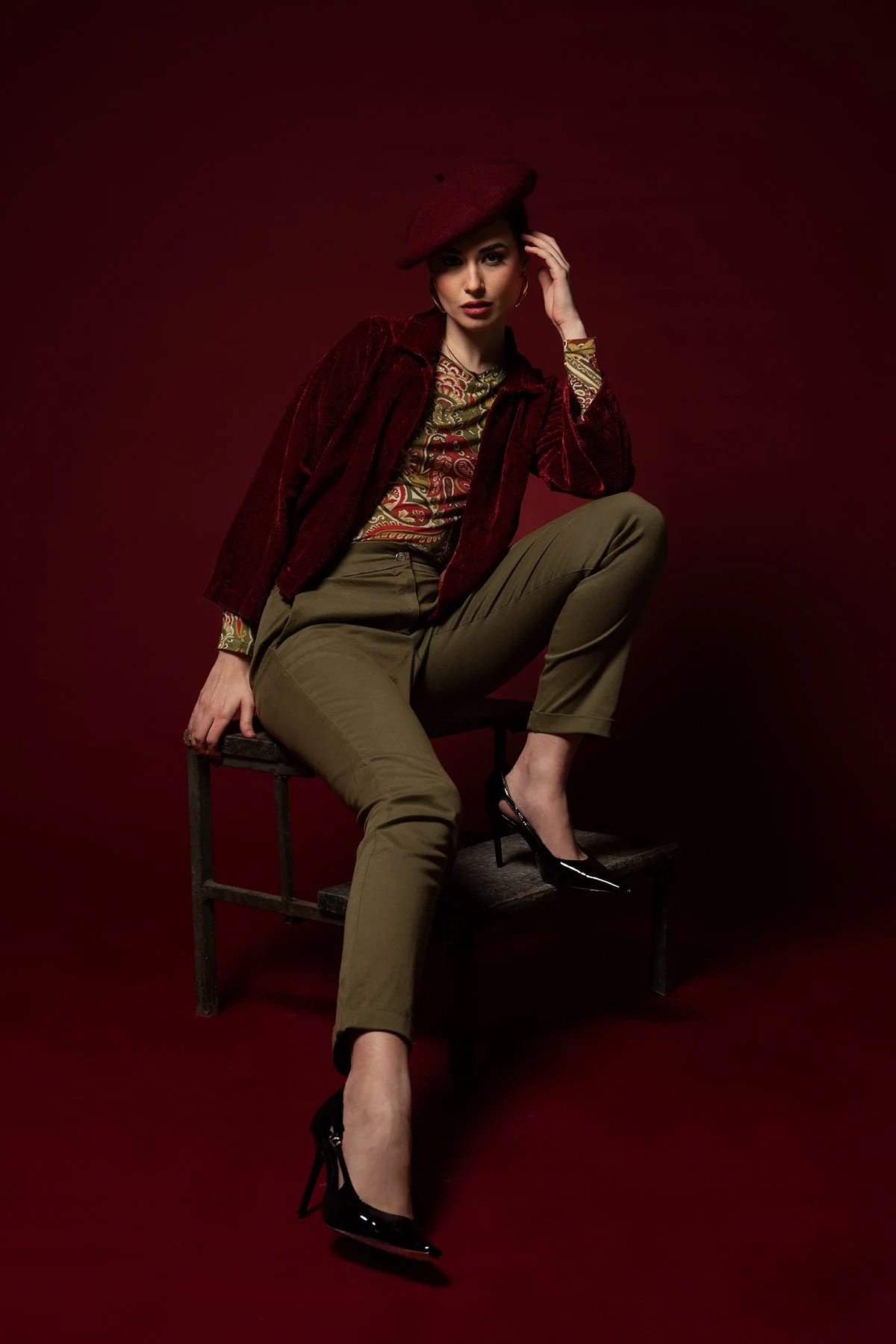 Donna seduta su una sedia con sfondo rosso scuro, indossa una giacca di velluto rosso, pantaloni kaki e tacchi neri, con un cappello rosso posato sulla testa.