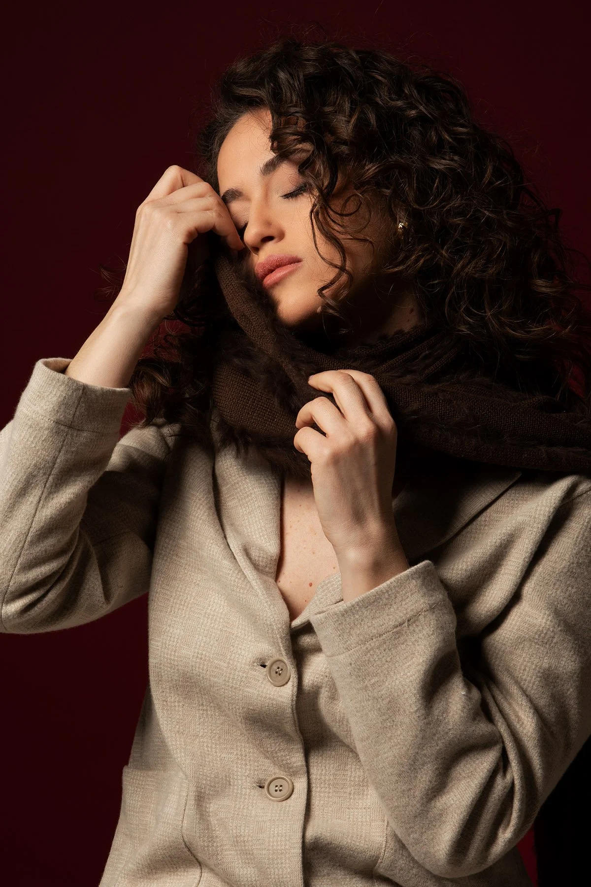 Una donna con capelli ricci, vestita con un cappotto beige, si trova in primo piano con uno sfondo rosso scuro, tiene una sciarpa intorno al collo e sembra pensierosa.