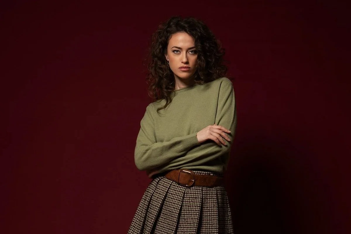 Donna con capelli ricci, indossa una maglia verde oliva e una gonna a pieghe con cintura marrone, sfondo rosso scuro.