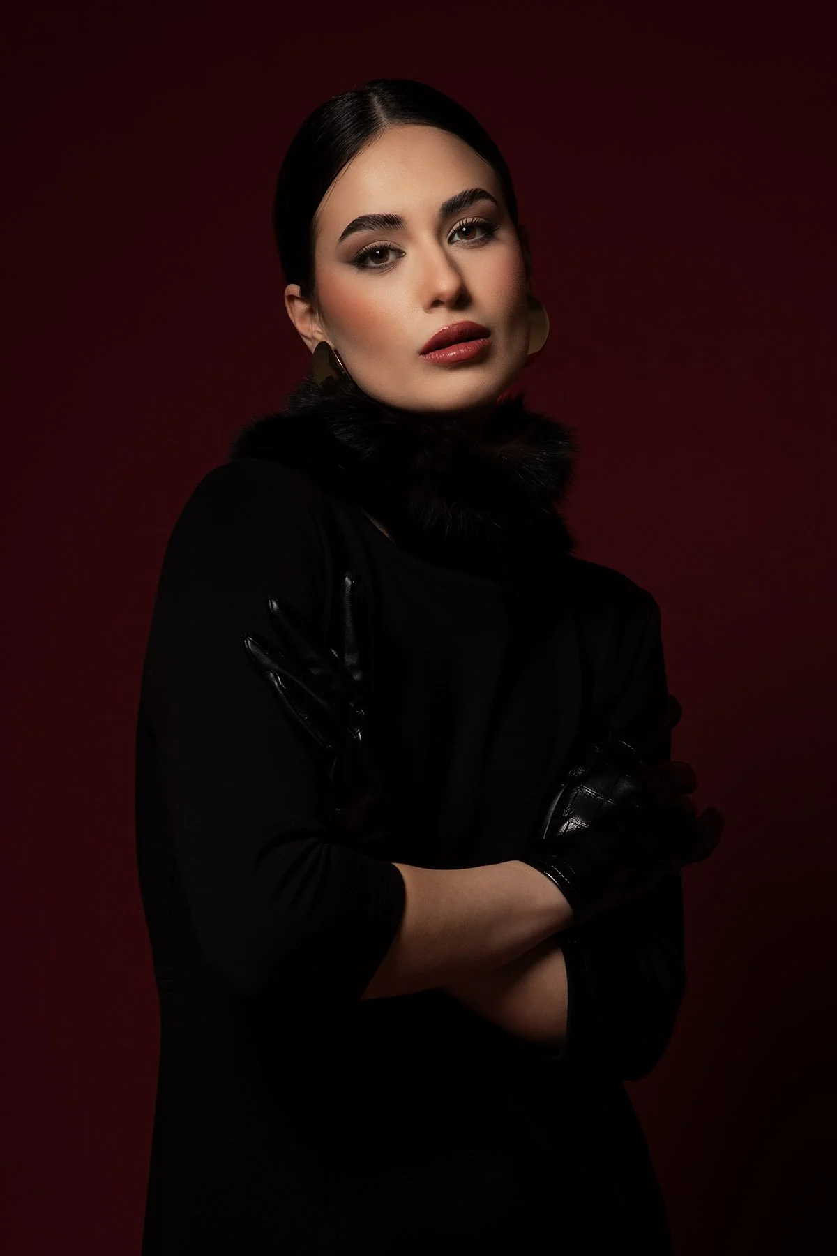 Ritratto di una donna con sopracciglia folte, trucco elegante e capelli scuri raccolti, indossa un cappotto nero con pelliccia e guanti in pelle, sfondo rosso scuro.