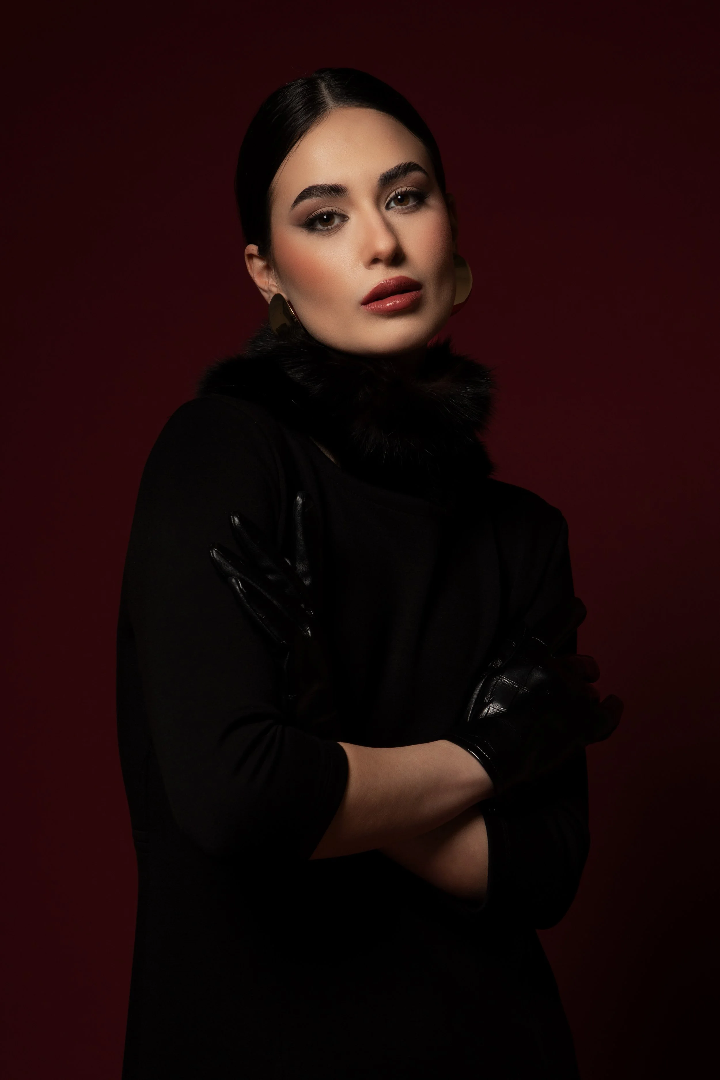 Ritratto di una donna con capelli castani scuri e trucco elegante, indossa un cappotto nero con pelliccia e orecchini grandi, sfondo sfumato rosso.