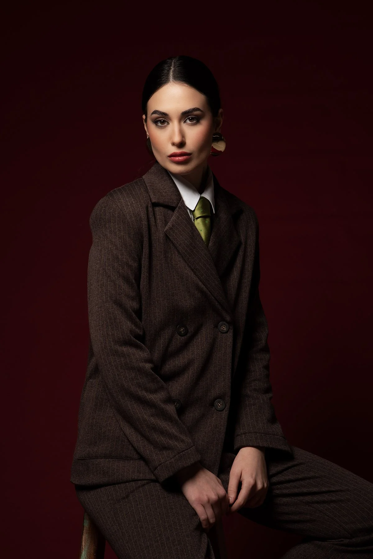 Una donna con capelli scuri, occhi intensi e trucco marcato, veste un completo doppiopetto marrone a quadri e una cravatta verde, su uno sfondo rosso scuro.