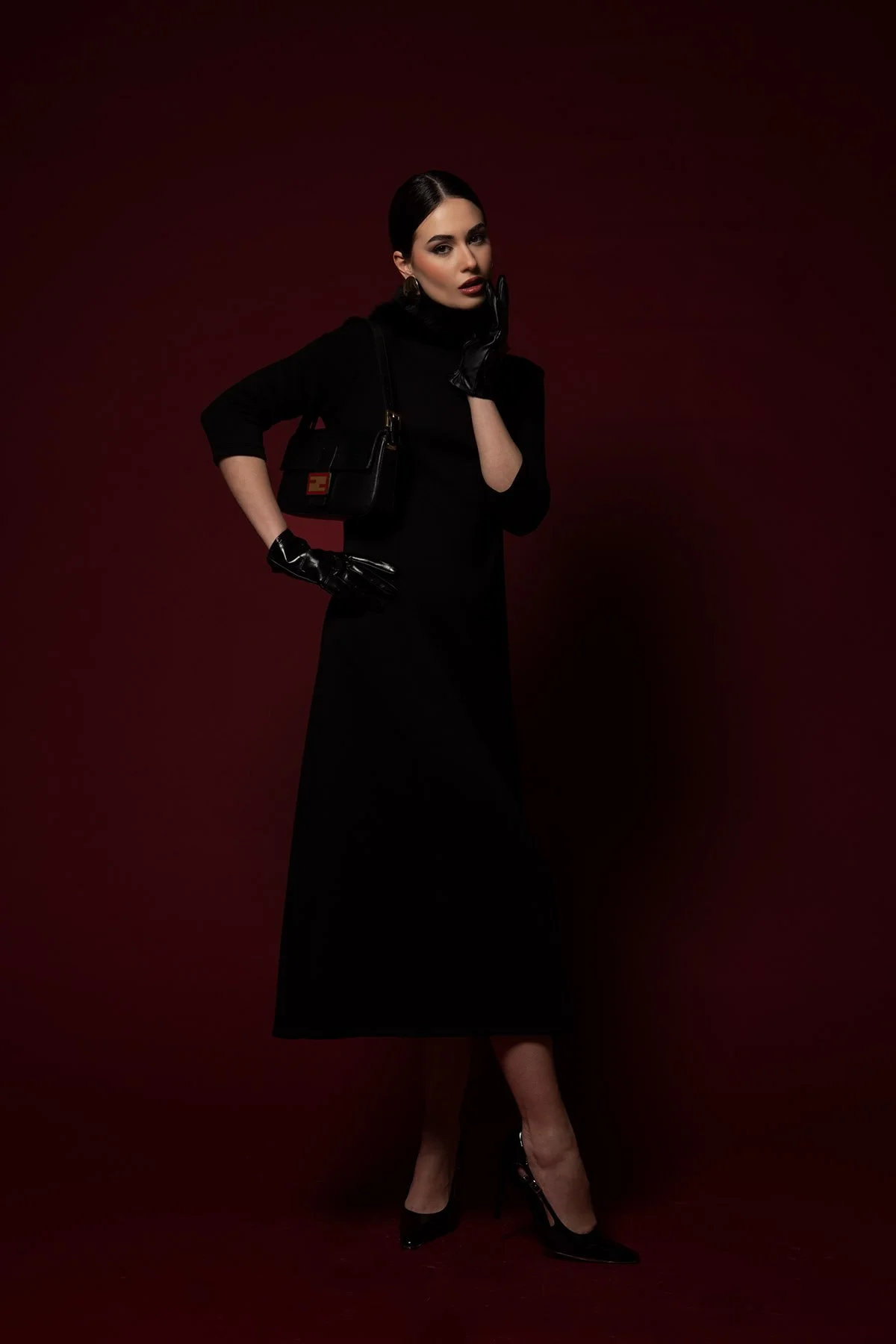 Donna in abito nero lungo, con occhiali e orecchini d'oro, indossa guanti in pelle nera, troviamo una borsa nera con chiusura dorata, sfondo sfumato rosso scuro.