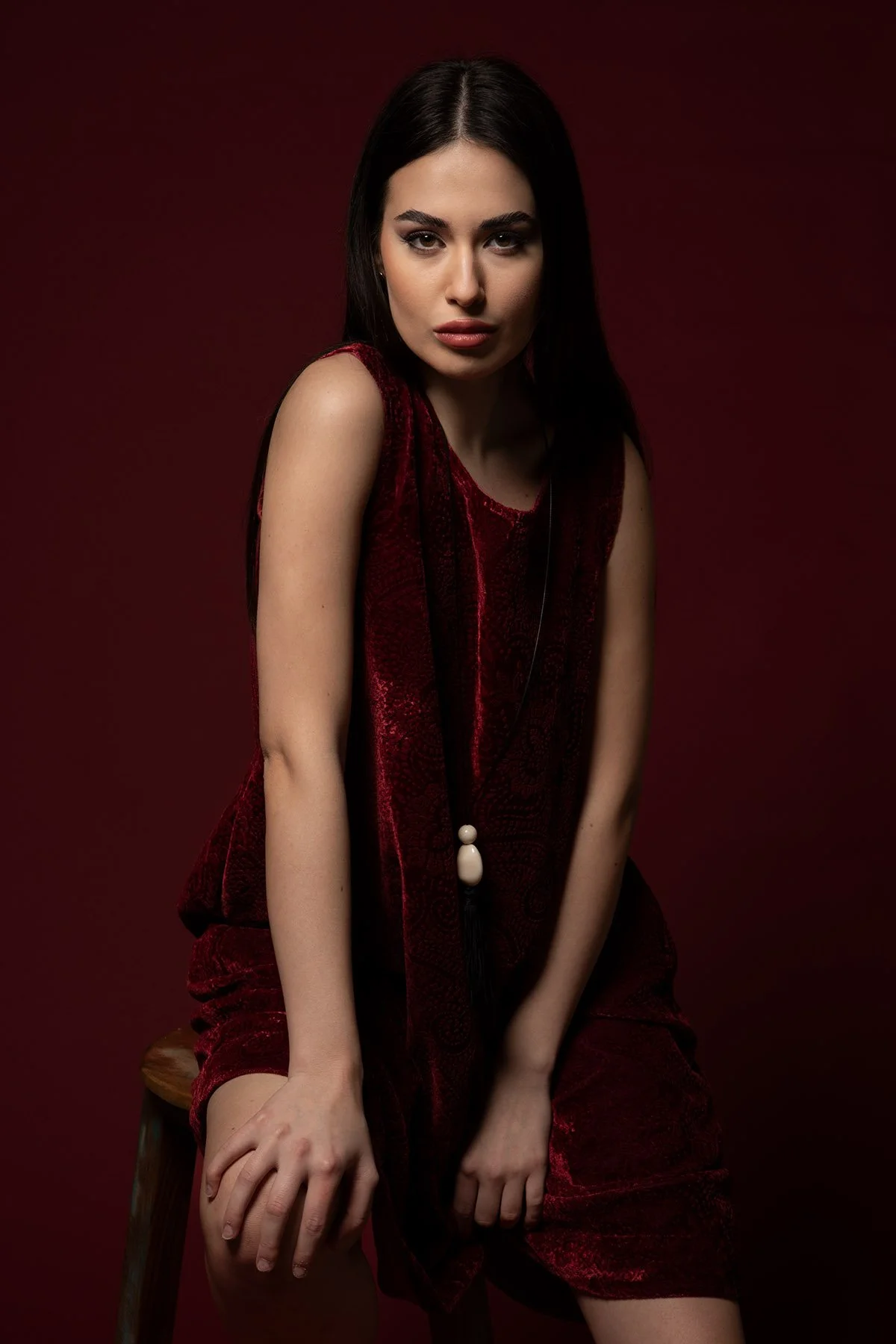 Ragazza con capelli scuri e vestito rosso seduta su uno sgabello contro uno sfondo rosso scuro.