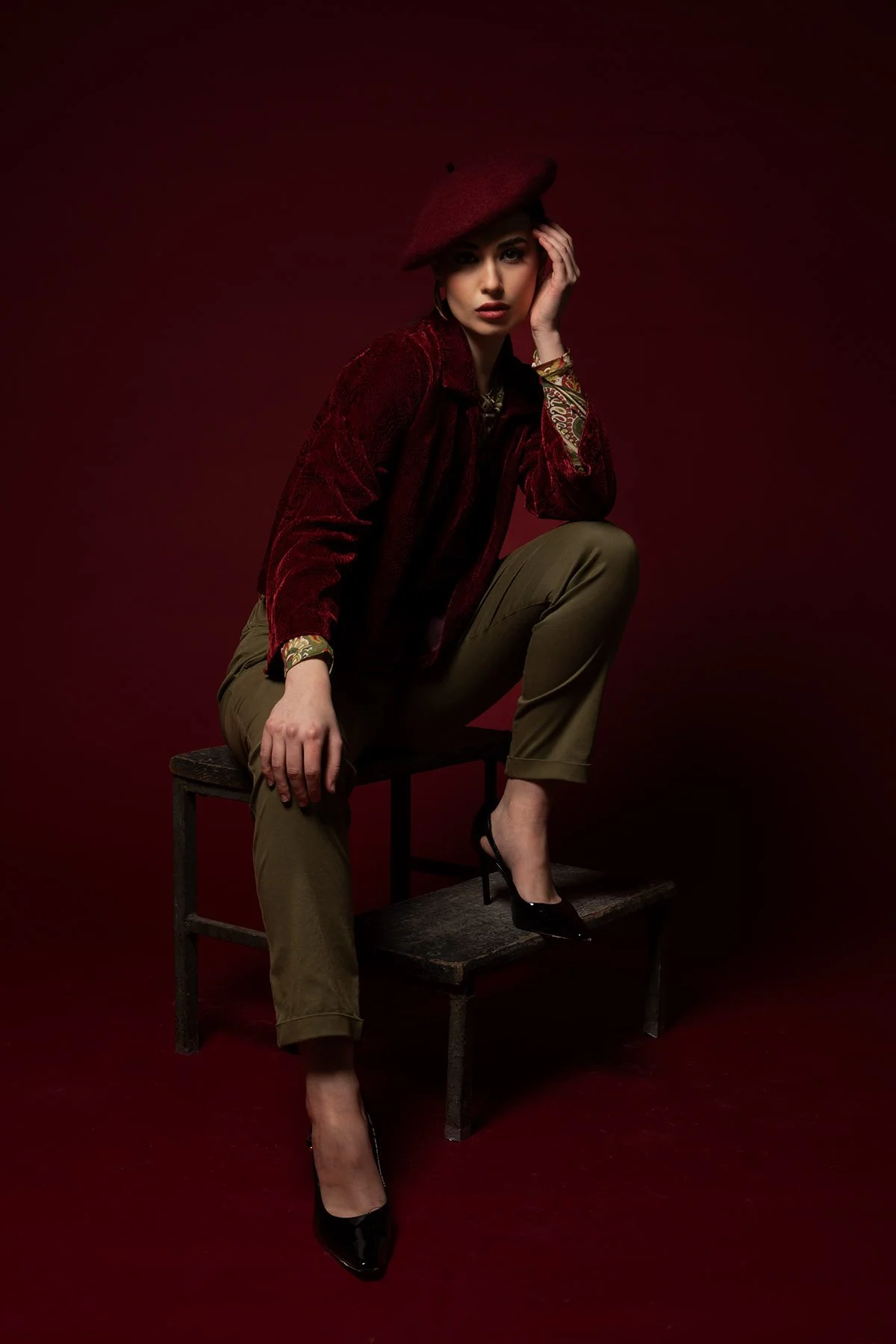 Una donna seduta su una sedia, indossa un cappello rosso e un abito rosso con pantaloni kaki e tacchi neri, sfondo scuro rosso.