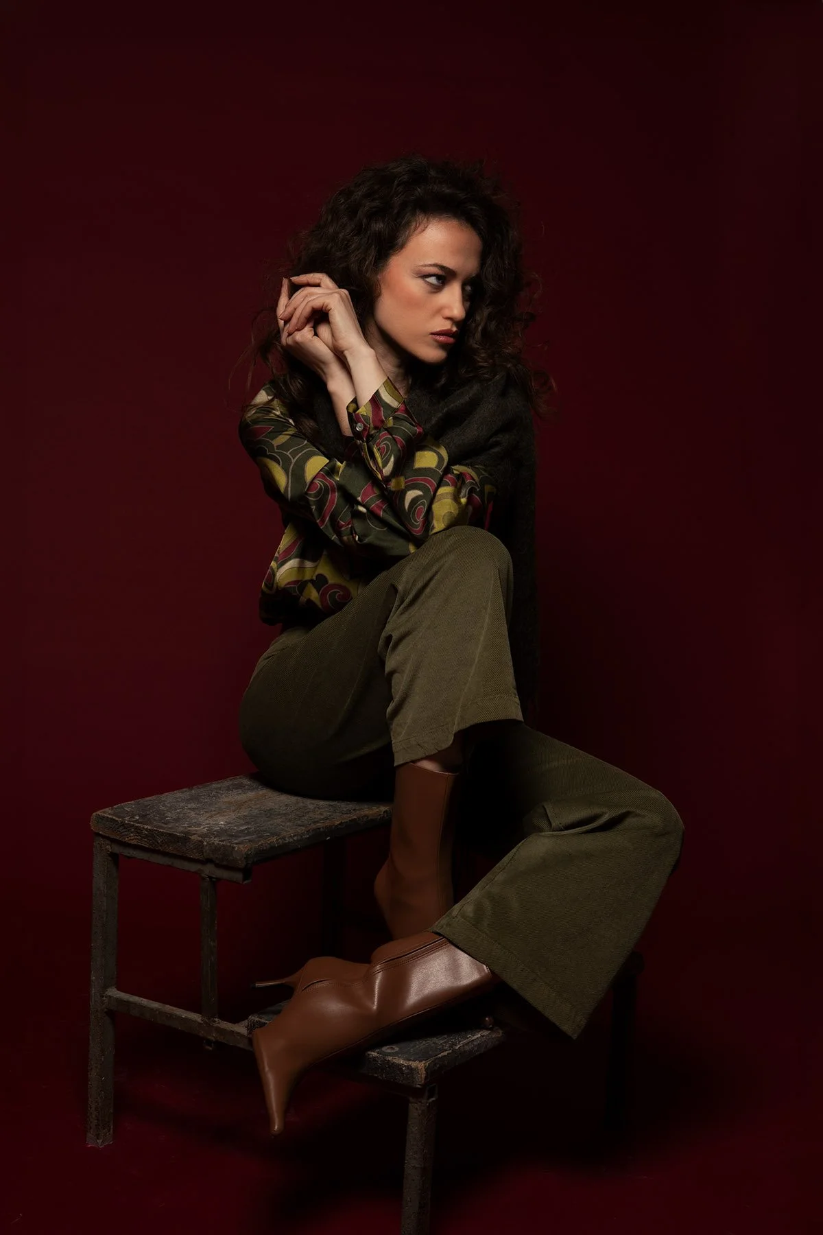 Donna seduta su uno sgabello di legno contro sfondo rosso scuro, vestita con pantaloni verdi, stivaloni marroni e camicetta con disegni geometrici.