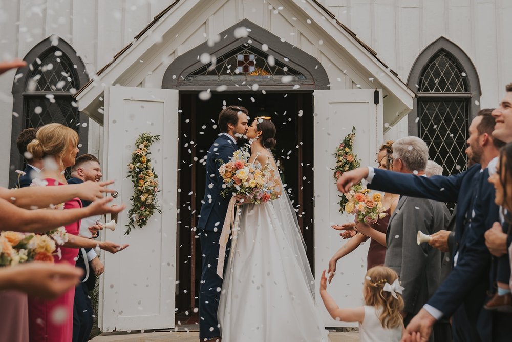 Wedding guests throwing petals over bride and groom outside chapel