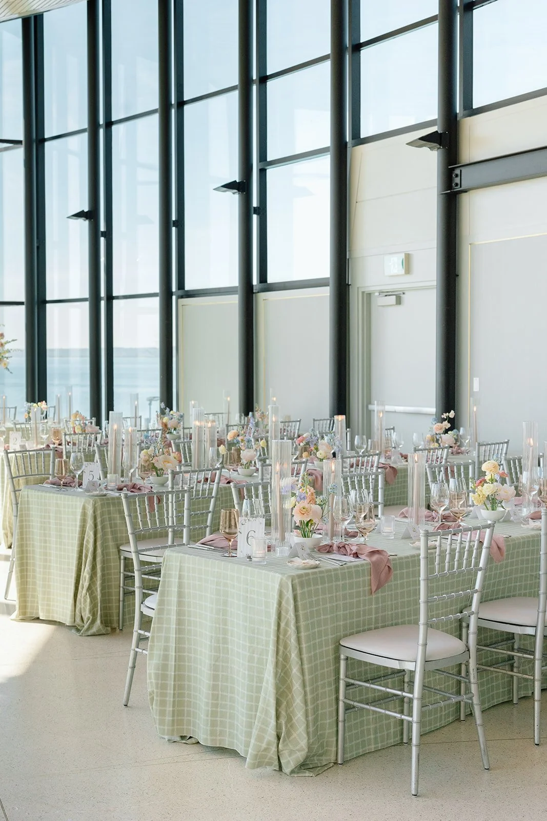  The magic of twilight ambient lighting. Long estate tables feature an elegant repetition of tall, fluted glass hurricane sleeves with glowing taper candles, casting a romantic reflection against the waterfront glass windows. Captured by Paula Visco 