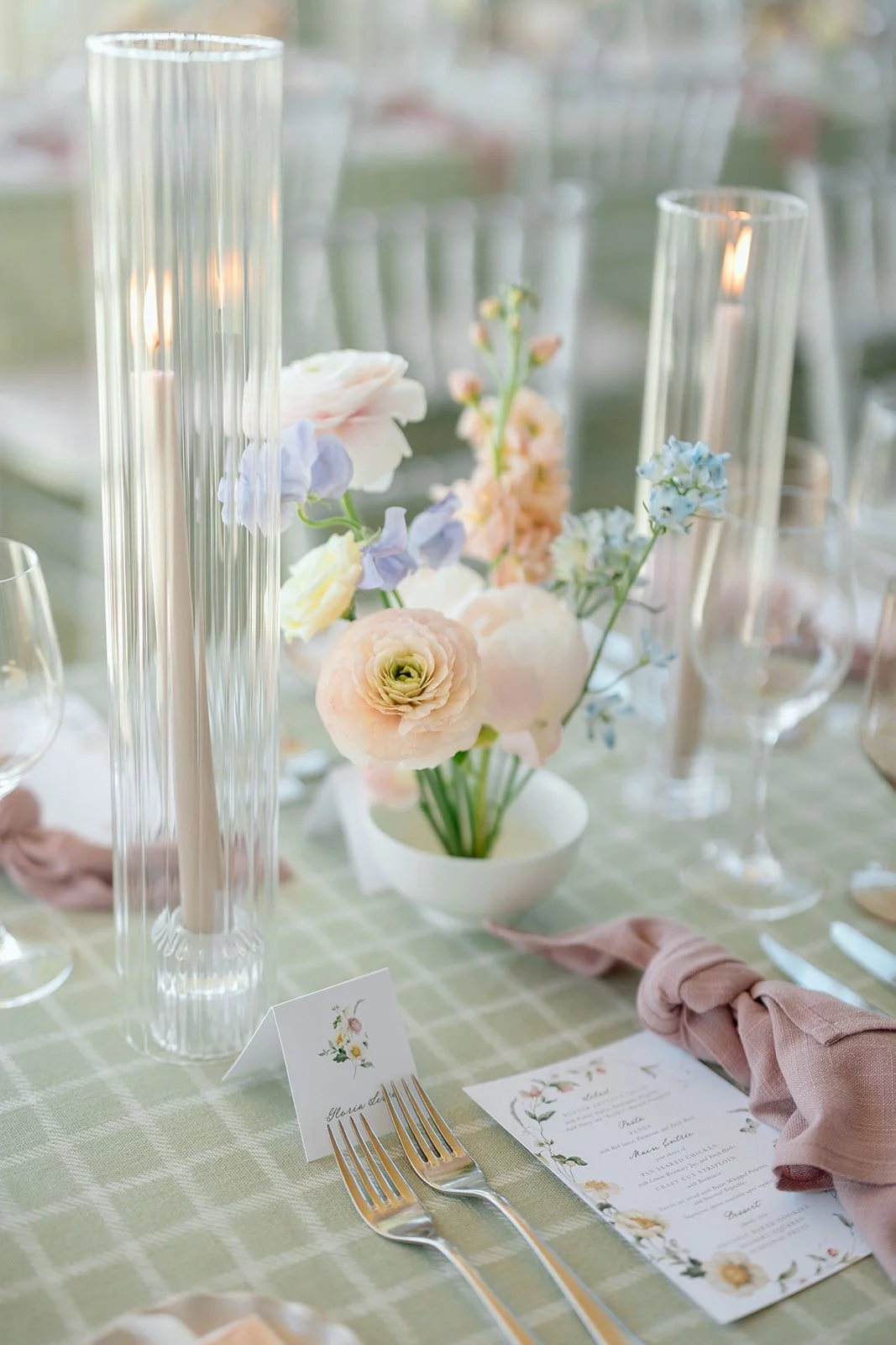  Close-up of our bespoke tablescape design. We paired a contemporary sage green checkered textile with an effortlessly knotted mauve linen napkin. A custom botanical menu and delicate fluted glass candle pillars complete this highly textured, modern-