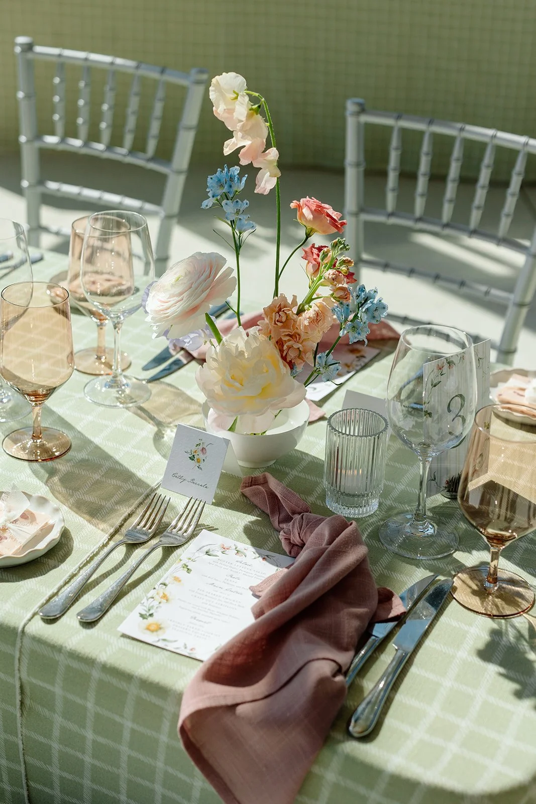  Low-profile white ceramic bowls were filled with airy, romantic arrangements of soft pink ranunculus, open white peonies, and playful stems of sky-blue delphinium. Elevating the main tables, grand white classical urns mimicked the light-filled grand