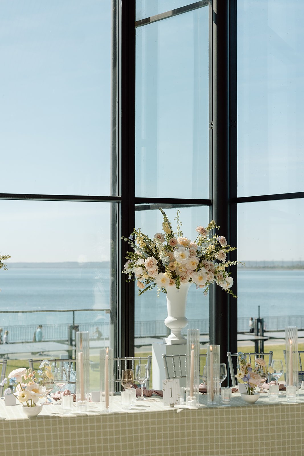  Large pastel floral urn arrangement featuring blush garden roses, peach ranunculus, and light blue delphinium on a white pedestal in front of Lake Ontario waterfront windows. 