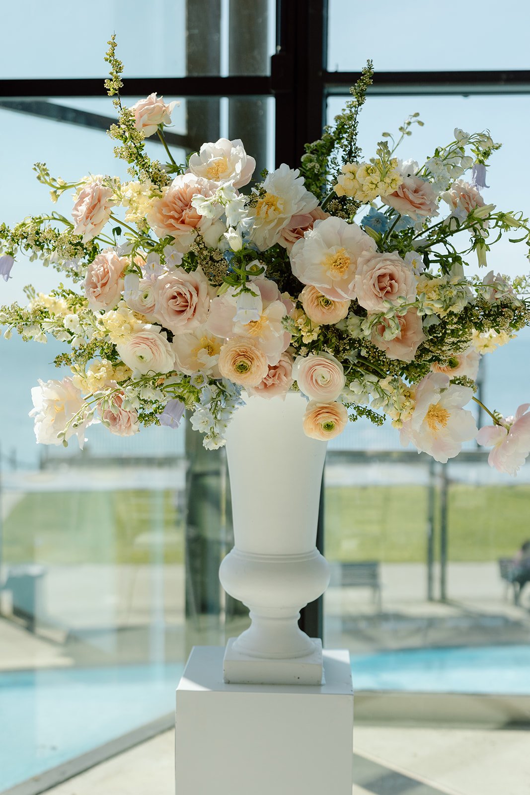  Statement floral design at its finest. This elevated white urn arrangement anchors the head table, utilizing airy, unstructured delphinium, peonies, and garden roses to mimic the natural movement of the waterfront horizon outside. 