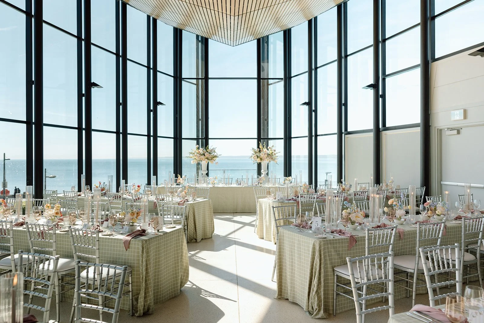  An architectural wide shot of the sun-filled event space at Spencer's at the Waterfront, showcasing the seamless flow of silver chiavari chairs and custom sage checkered linens against the lake. 