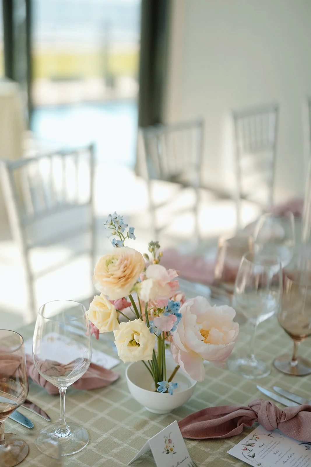 Low-profile white ceramic bowls were filled with airy, romantic arrangements of soft pink ranunculus, open white peonies, and playful stems of sky-blue delphinium. Elevating the main tables, grand white classical urns mimicked the light-filled grand