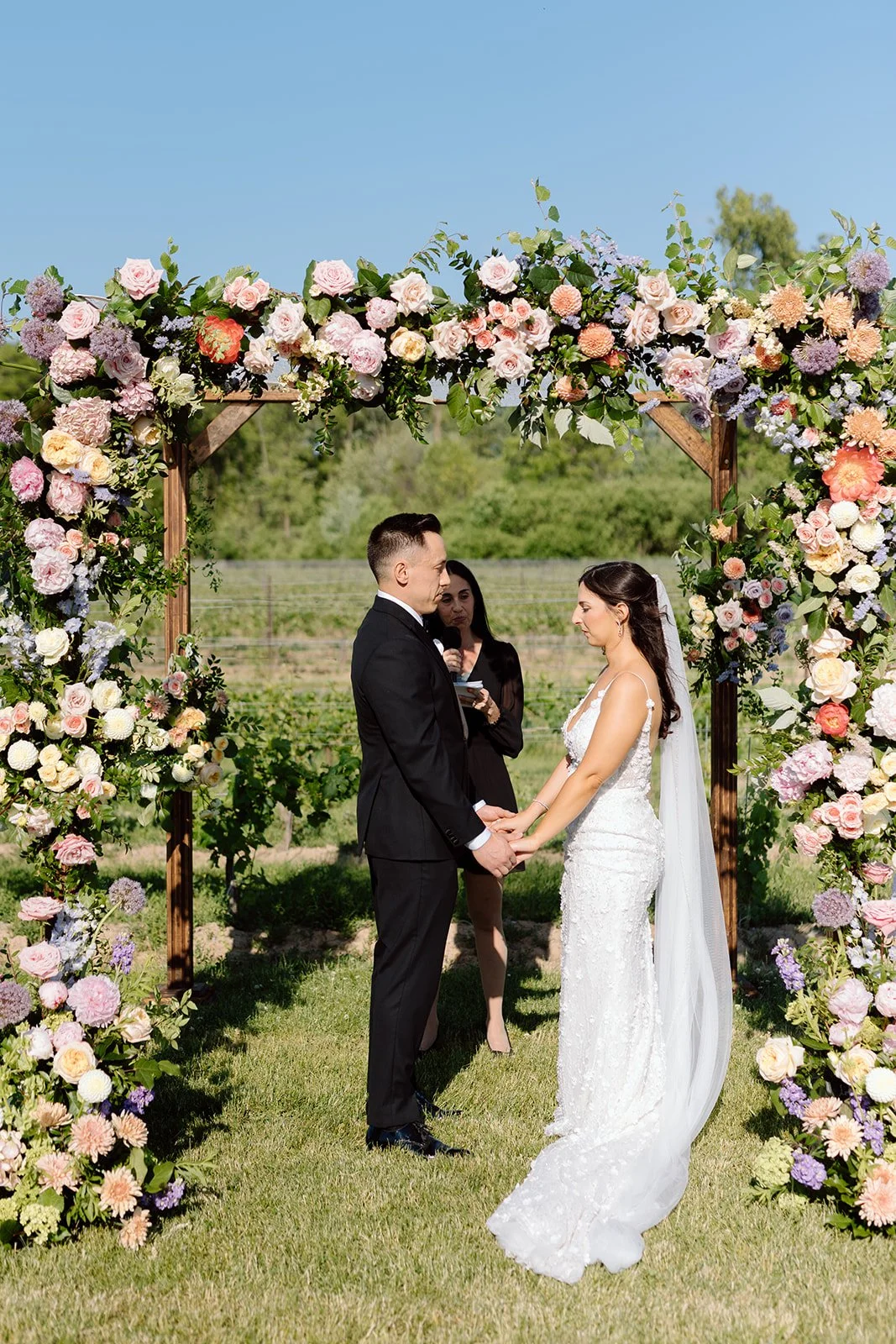   Alt Text:  Summer vineyard wedding ceremony featuring an asymmetrical floral chuppah with coral, peach, lavender, and cream garden flowers under a clear blue sky. 