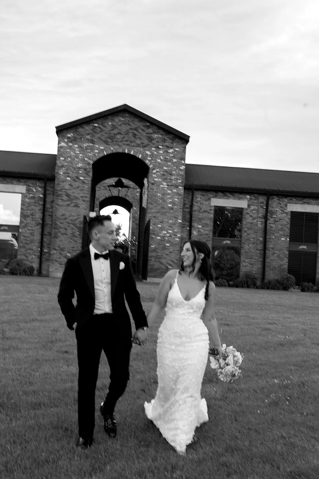  lack and white sunset wedding portrait of a bride and groom walking on the lawn in front of the brick estate at The Hare Wine Co. 