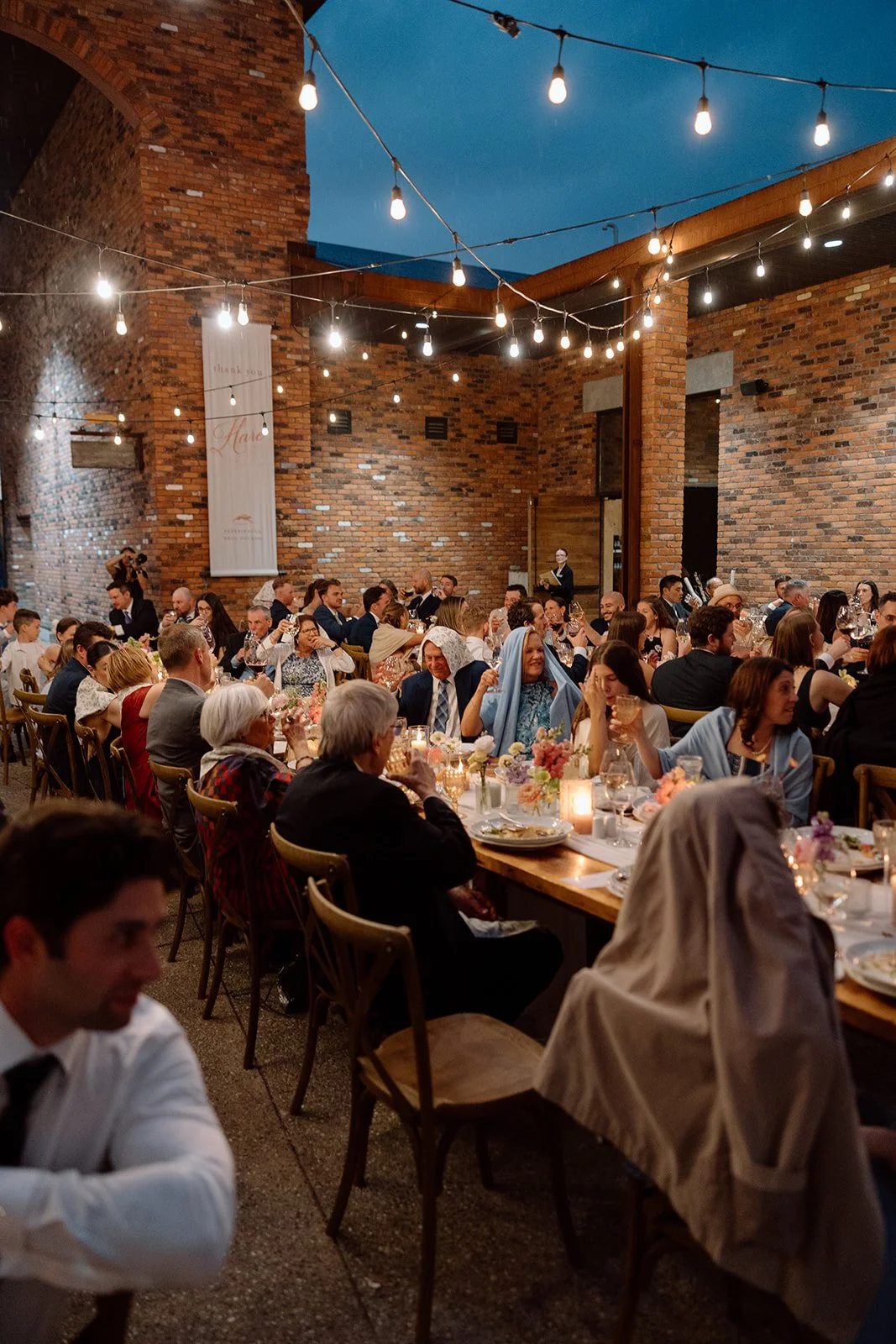  Intimate open-air winery courtyard reception layout featuring crossback chairs, candlelight, and glowing market string lighting. 