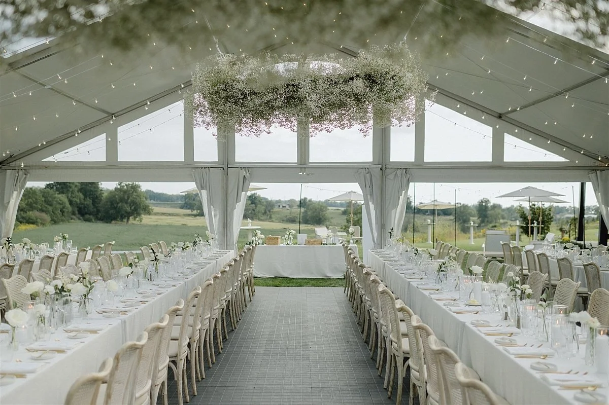 A wide photo of a white tented wedding reception area with a large floral installation hanging from the top of the tent