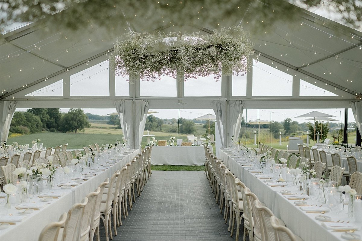 A wide photo of a white tented wedding reception area with a large floral installation hanging from the top of the tent