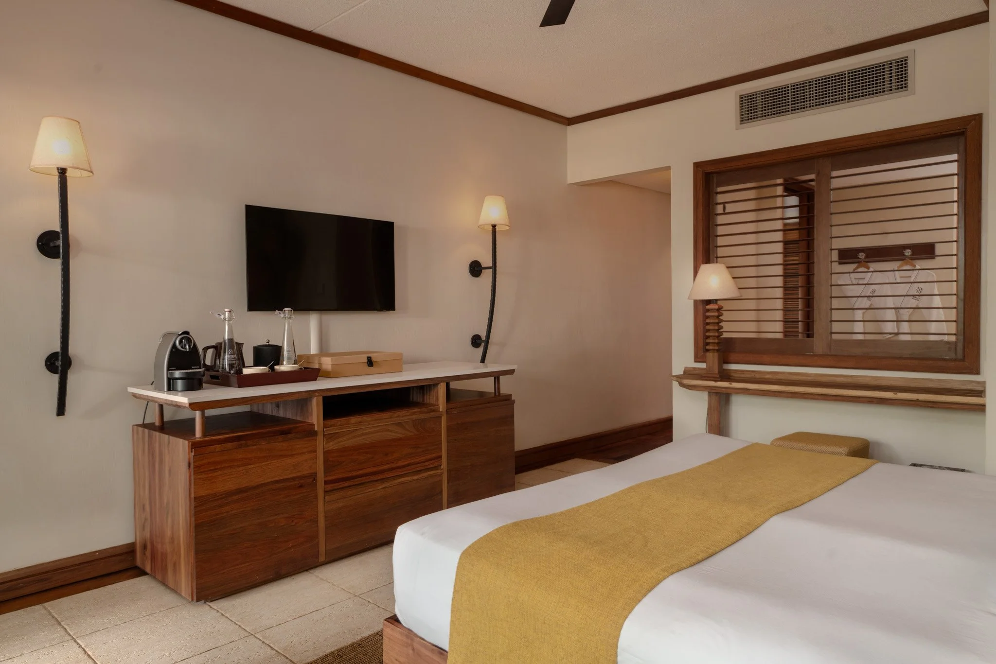 Hotel room with a bed, yellow bed runner, wooden furniture, wall-mounted lights, a TV, a window with wooden blinds, and a closet with robes.