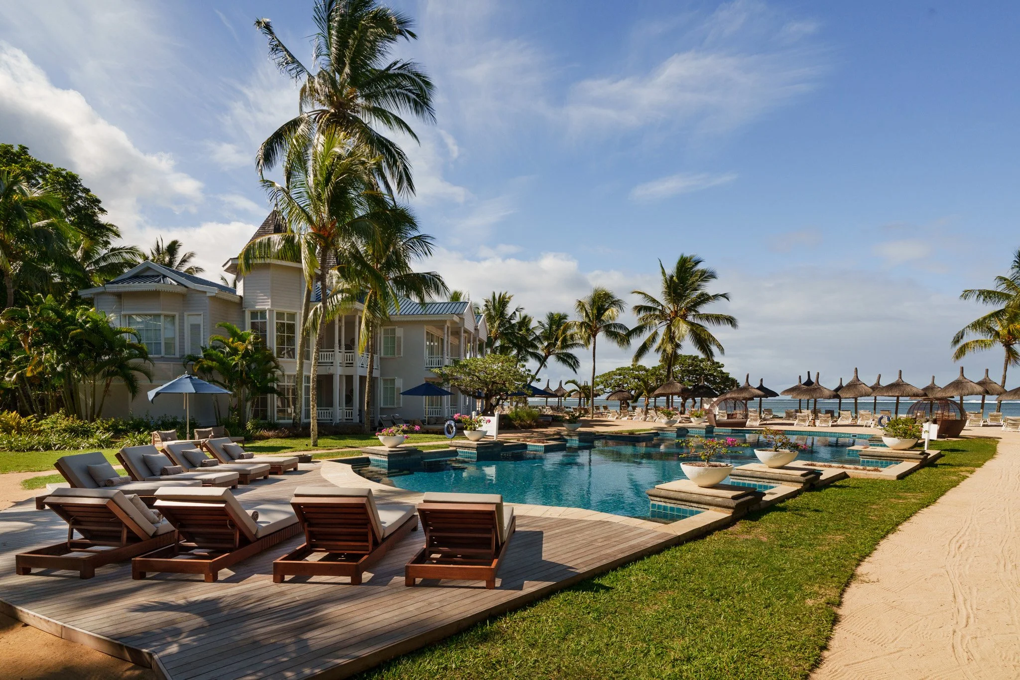 luxurious beachfront resort with a swimming pool, numerous sun loungers, palm trees, and thatched umbrellas along a sandy beach with ocean in the background