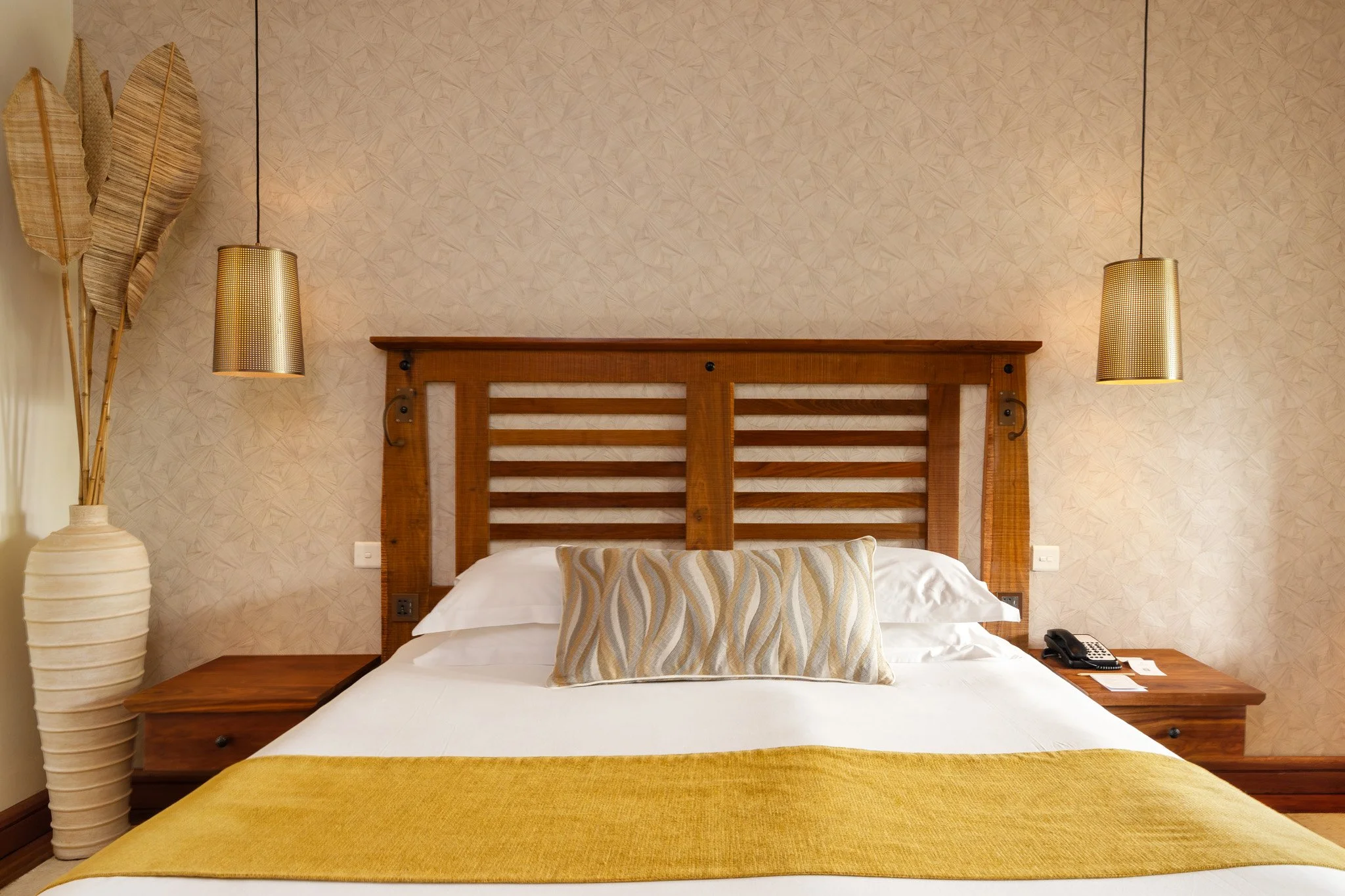 A neatly made bed with a wooden headboard, a decorative pillow, and a yellow bed runner. Two pendant lights hang from the ceiling on either side, and a large ceramic vase with tall dried leaves is placed on the left. A telephone and other small items