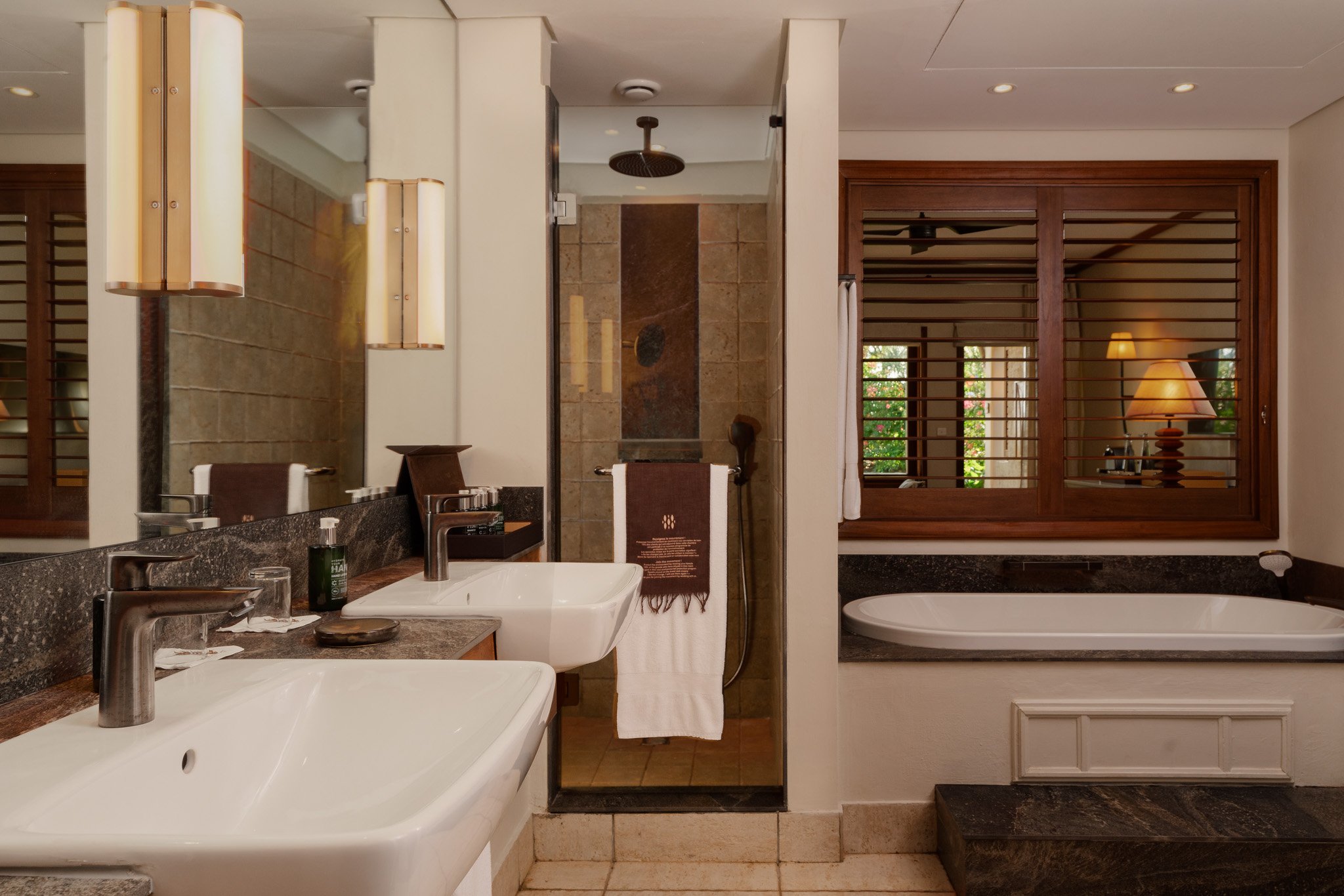 A luxurious bathroom with double sinks, a walk-in shower, a bathtub, and large windows with wooden shutters, decorated with warm lighting.
