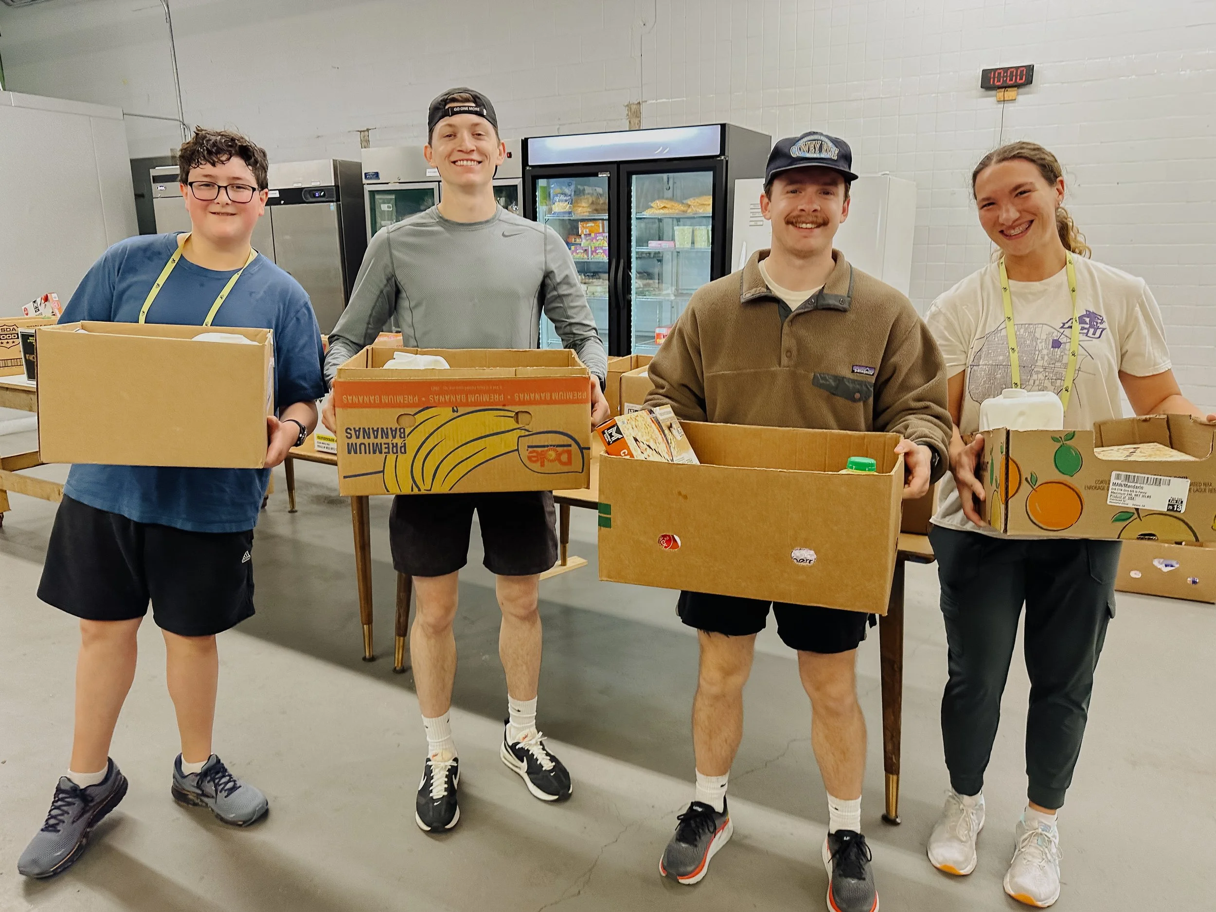 Four volunteers standing in a food bank with boxes of food, smiling at the camera.