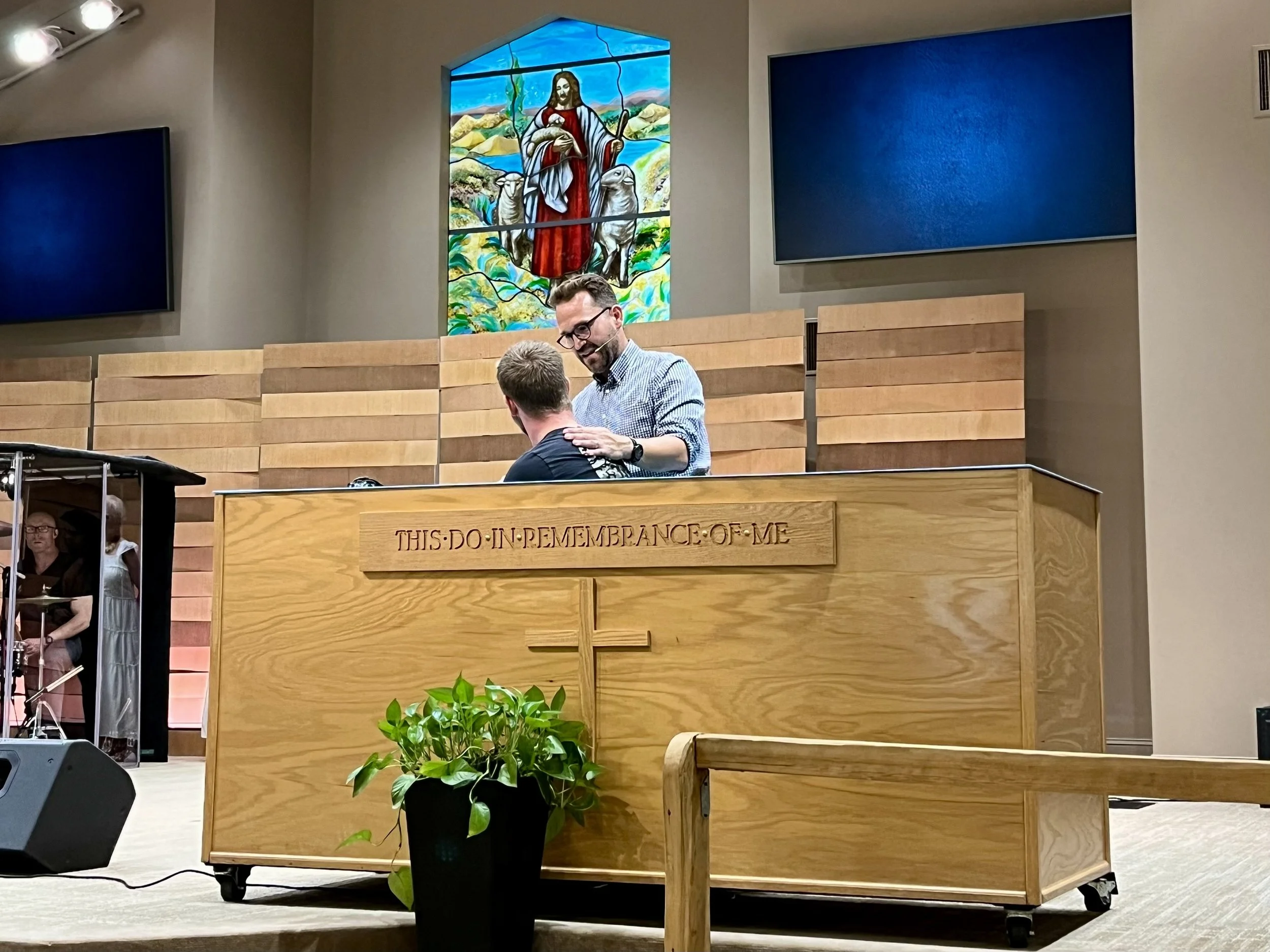 A man kneeling and praying on a wooden altar with a cross, inside a church, with another man standing and placing his hand on the kneeling man's shoulder. The altar has a plaque that reads 'THIS DO IN REMEMBRANCE OF ME'. There is a potted plant in front of the altar, stained glass window depicting Jesus with sheep behind them, and two large monitors behind.