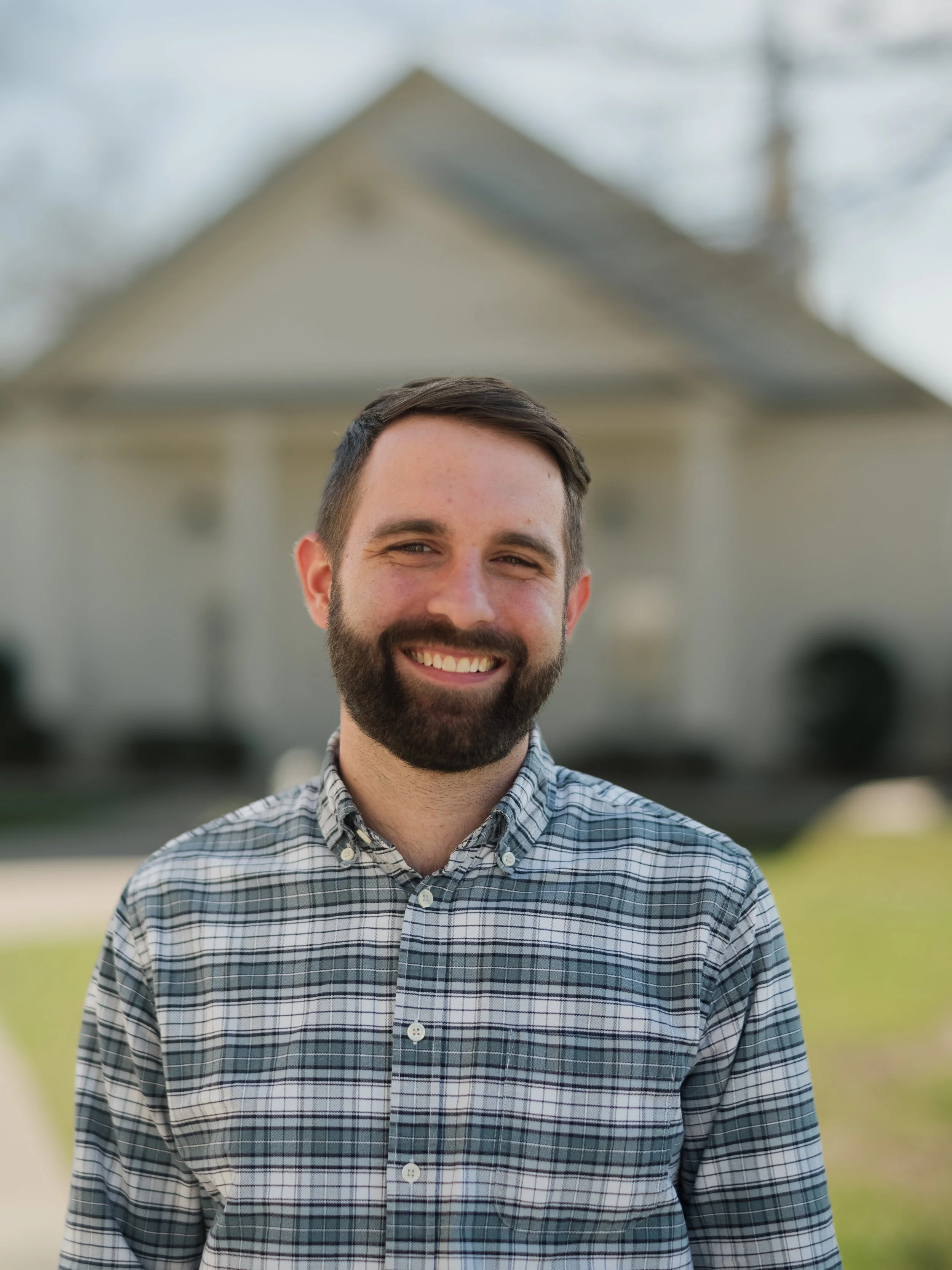 A man with dark hair, beard, and mustache wearing a plaid button-up shirt, smiling outdoors with a house in the background.