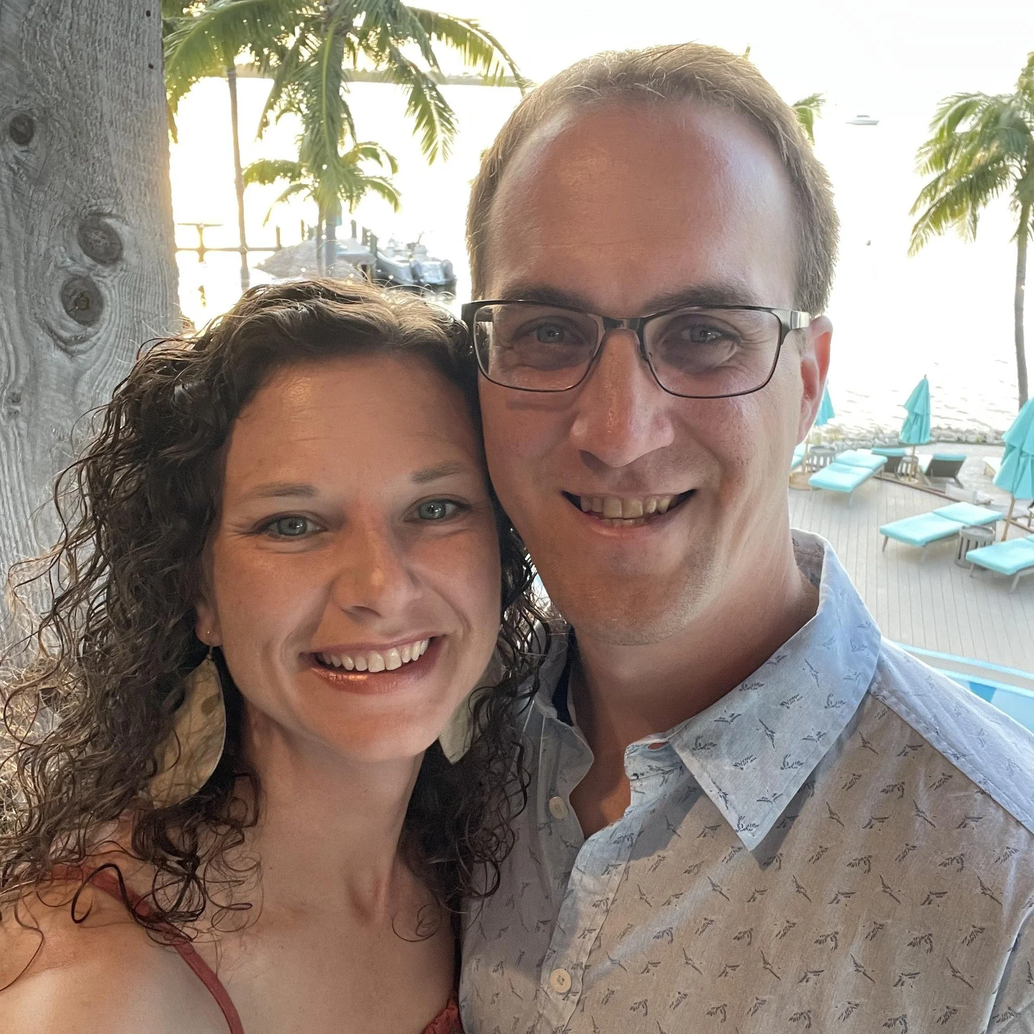 A smiling man and woman taking a selfie together on a beachside patio with palm trees, lounge chairs, and the ocean in the background.