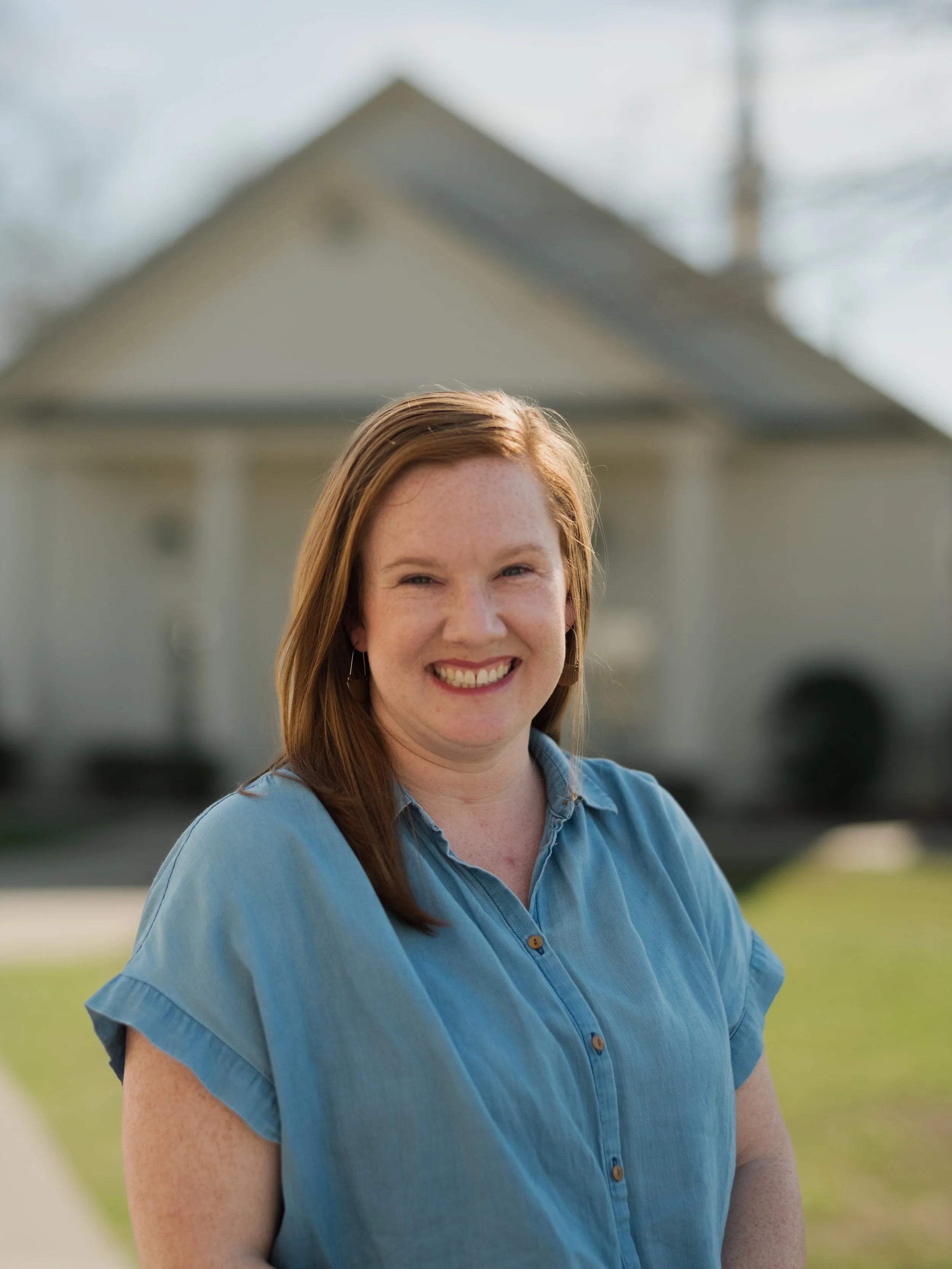 A smiling woman with red hair in a blue shirt standing outside a house.