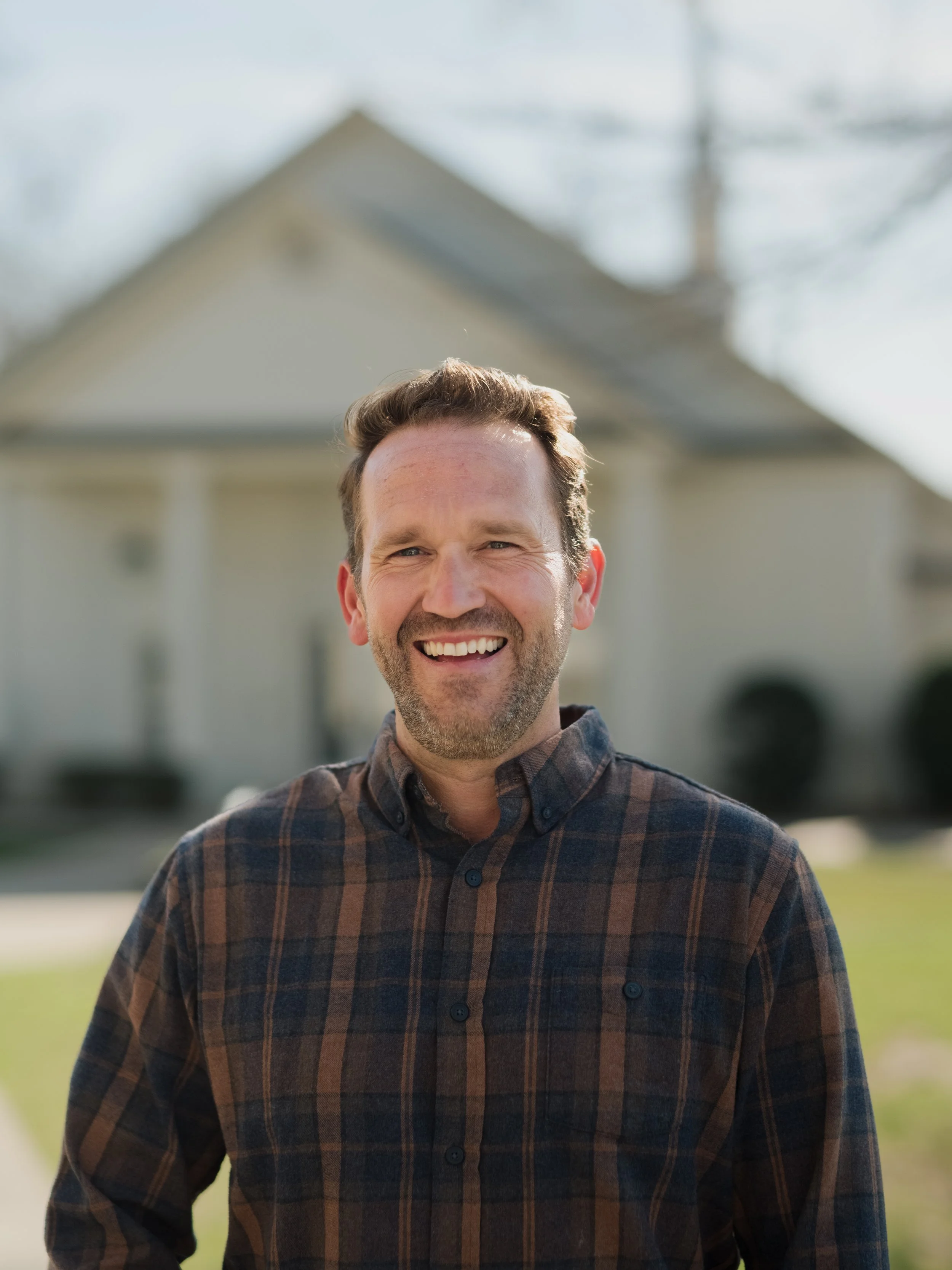 A man with short brown hair and a beard smiling outdoors in front of a house with a peaked roof during daytime.