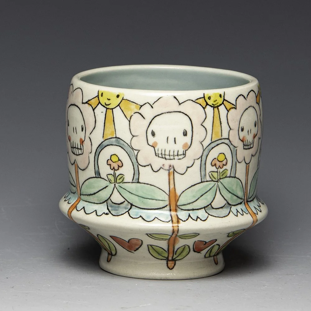 A ceramic cup with a colorful design featuring smiling skulls, flowers, and leaves, in a whimsical style.