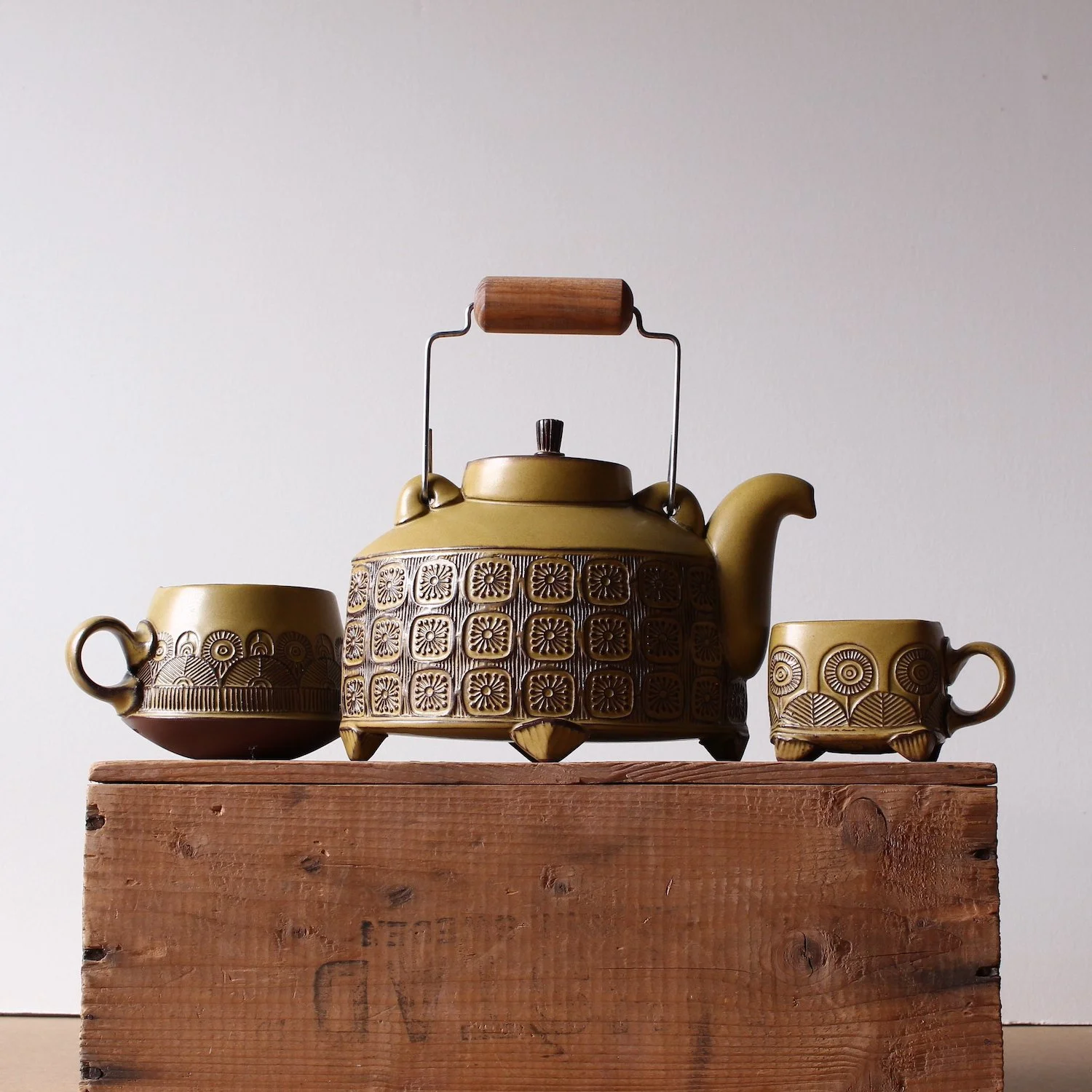 A decorative yellow teapot with intricate patterns, flanked by two matching cups, sits on a wooden box against a plain white background.