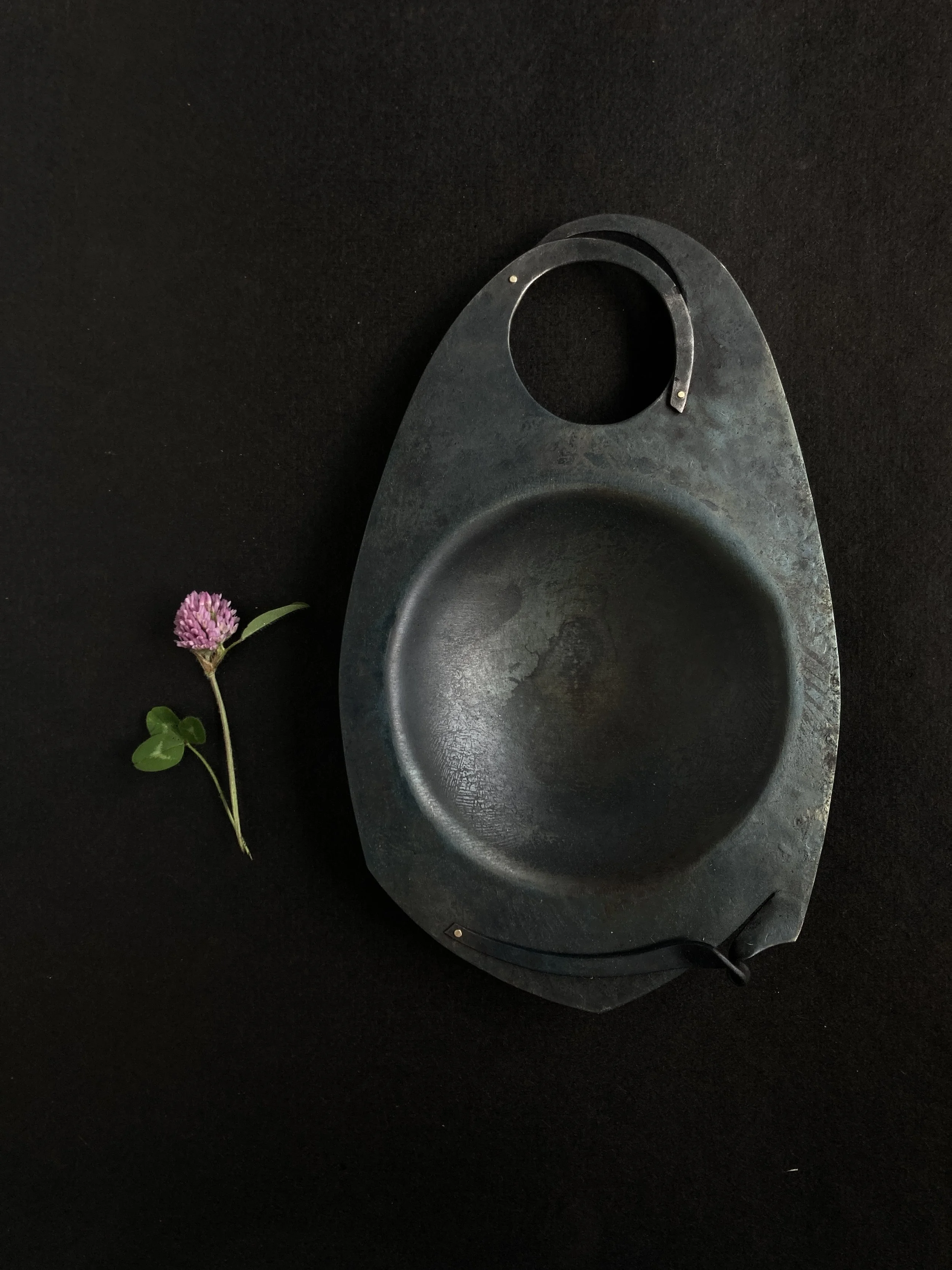 A worn cast iron skillet with a handle, placed on a dark surface, with a small pink flower and green leaves to the left of it.