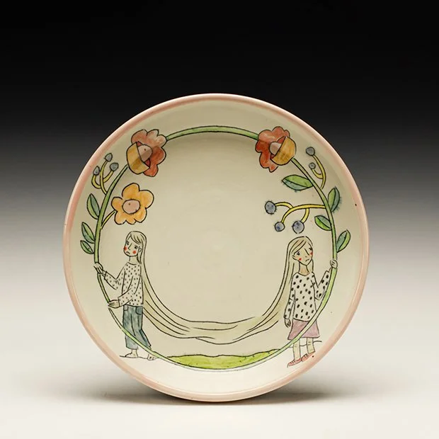 Decorative ceramic plate with illustrations of two girls holding long, flowing hair, surrounded by flowers and blueberry-like berries.