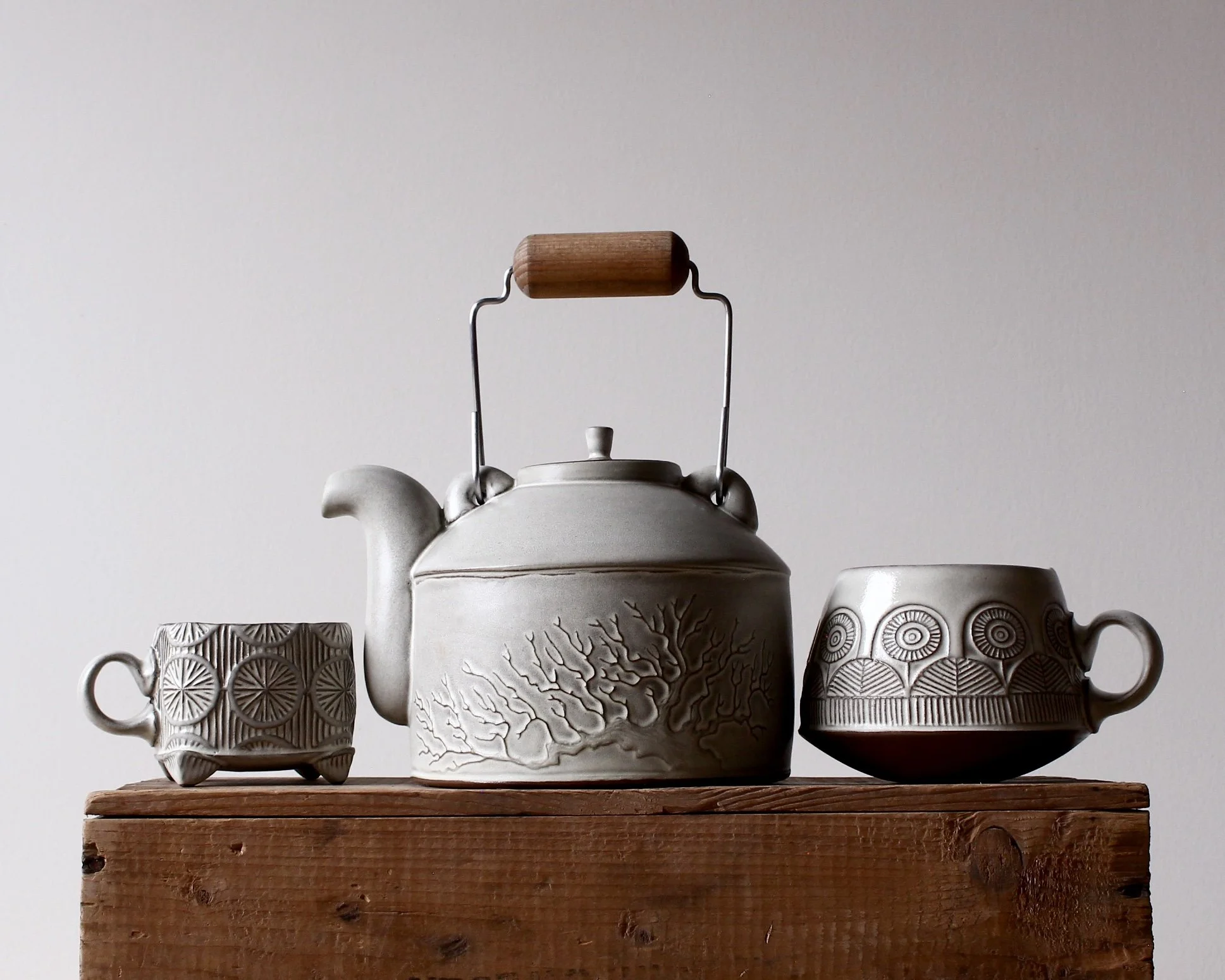 A ceramic teapot flanked by two ceramic mugs on a wooden surface against a plain background.