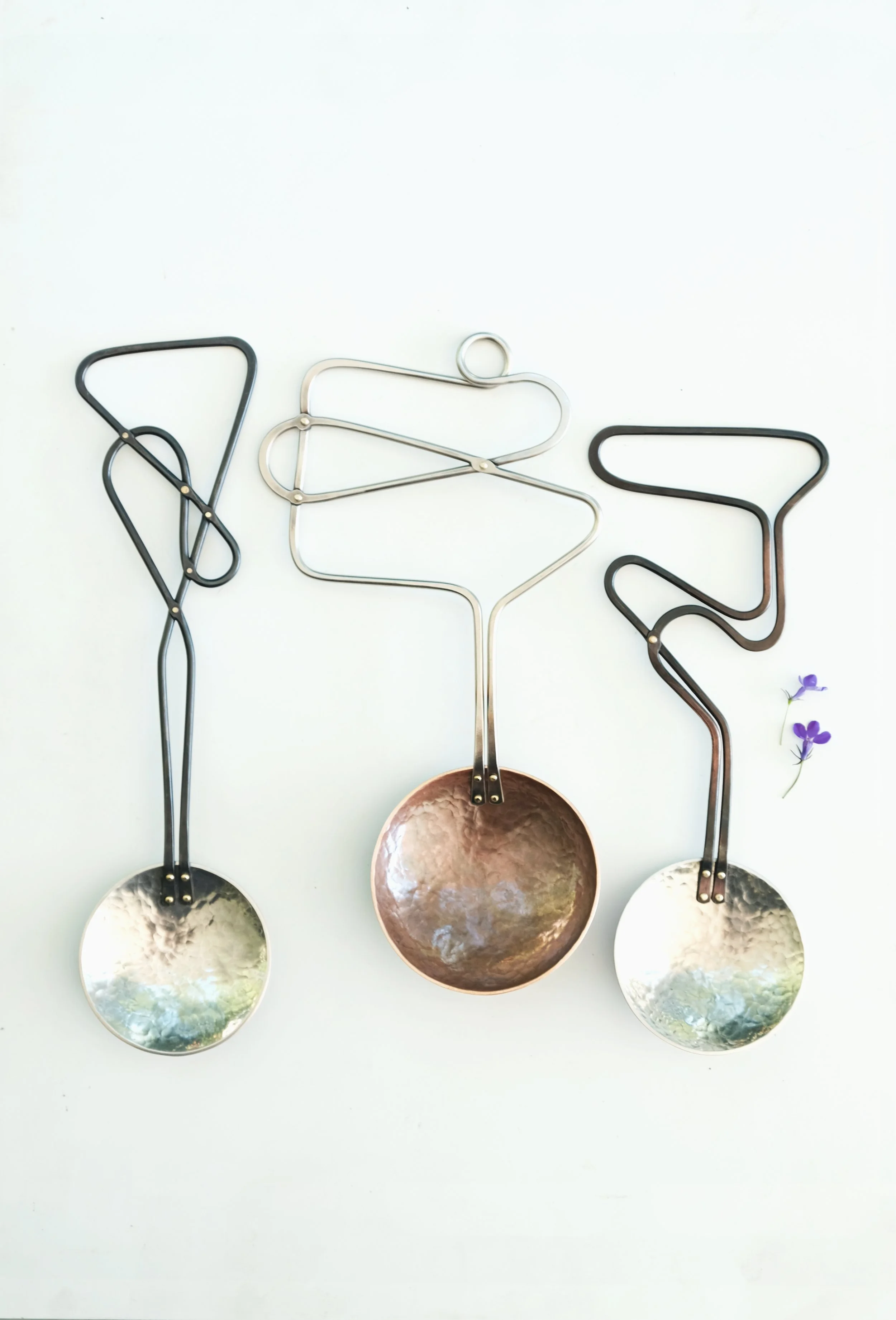 Three metal ladles with woven handles, arranged on a white surface with two small purple flowers in the corner.