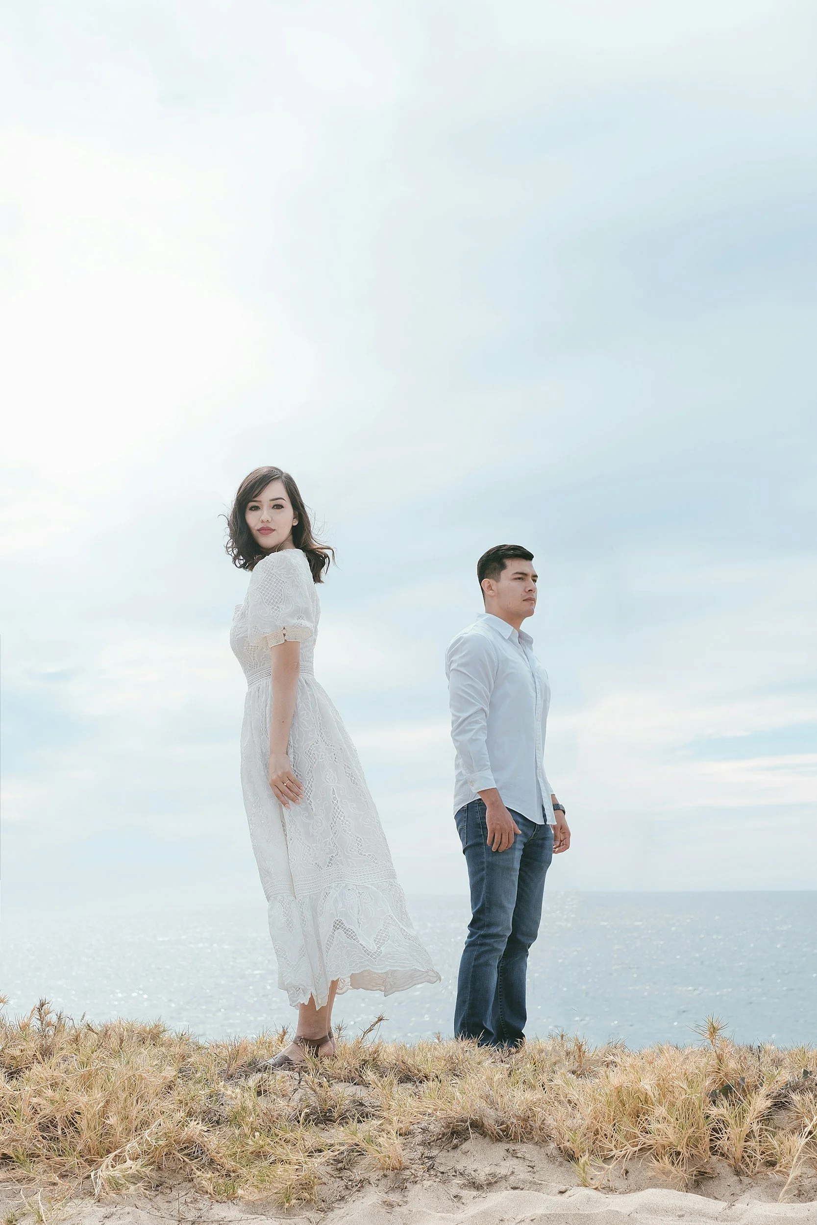 A young woman in a white dress and a young man in a light blue shirt and jeans standing on a sandy hill with grass, overlooking the ocean under a cloudy sky.