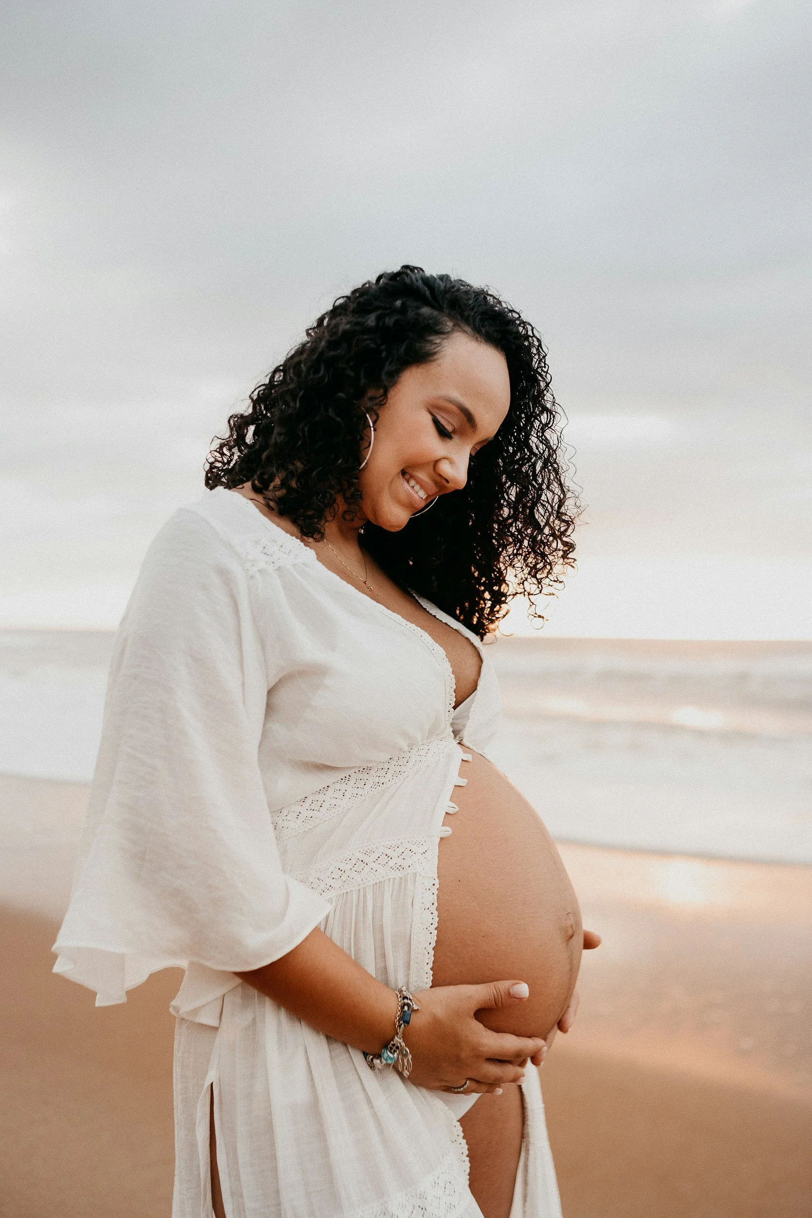 A pregnant woman with curly black hair wearing a white dress and jewelry, standing on a beach at sunset, smiling and gently holding her belly.
