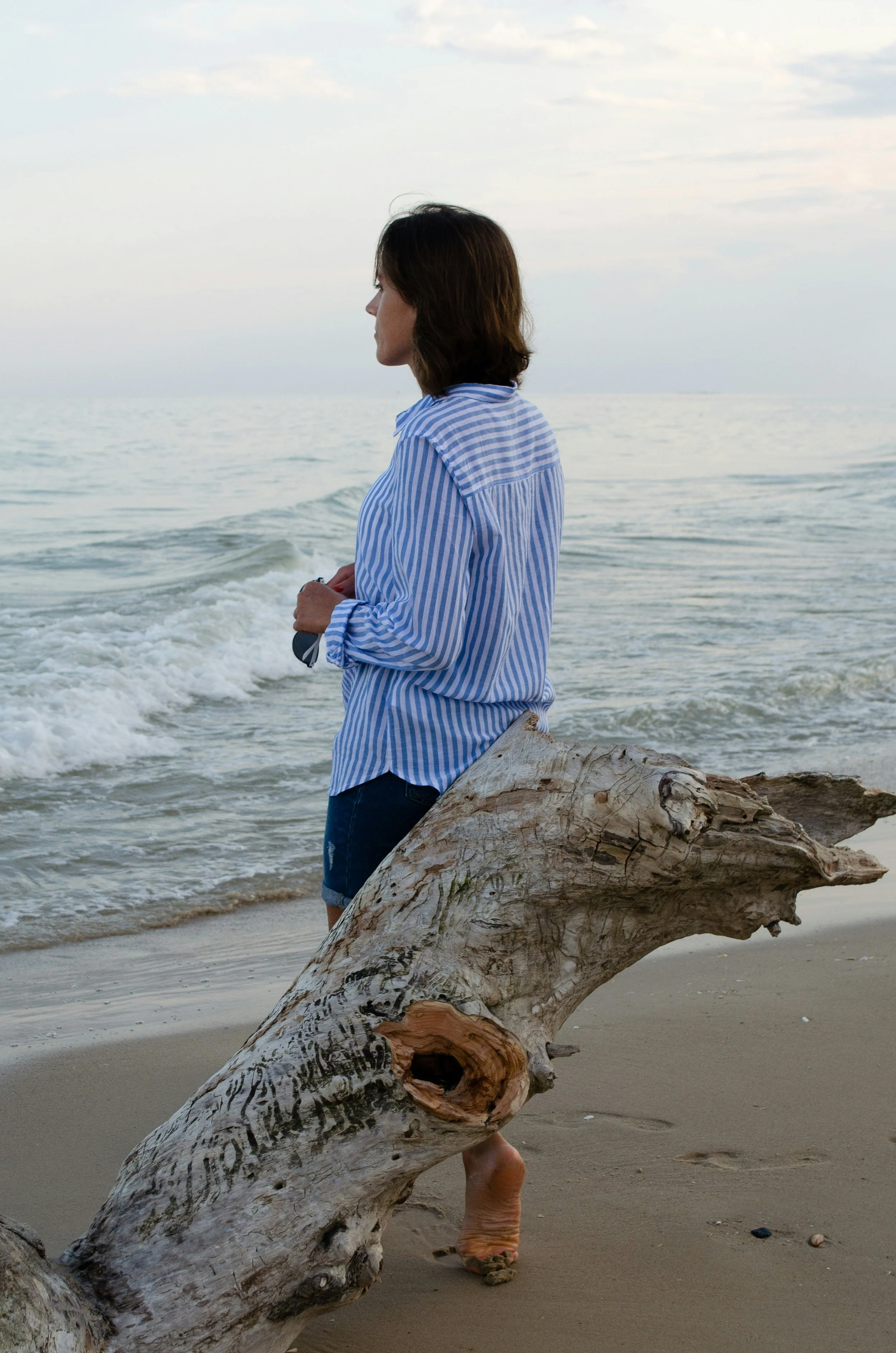 Woman in a blue and white striped shirt standing on a beach near a piece of driftwood, looking at the ocean.